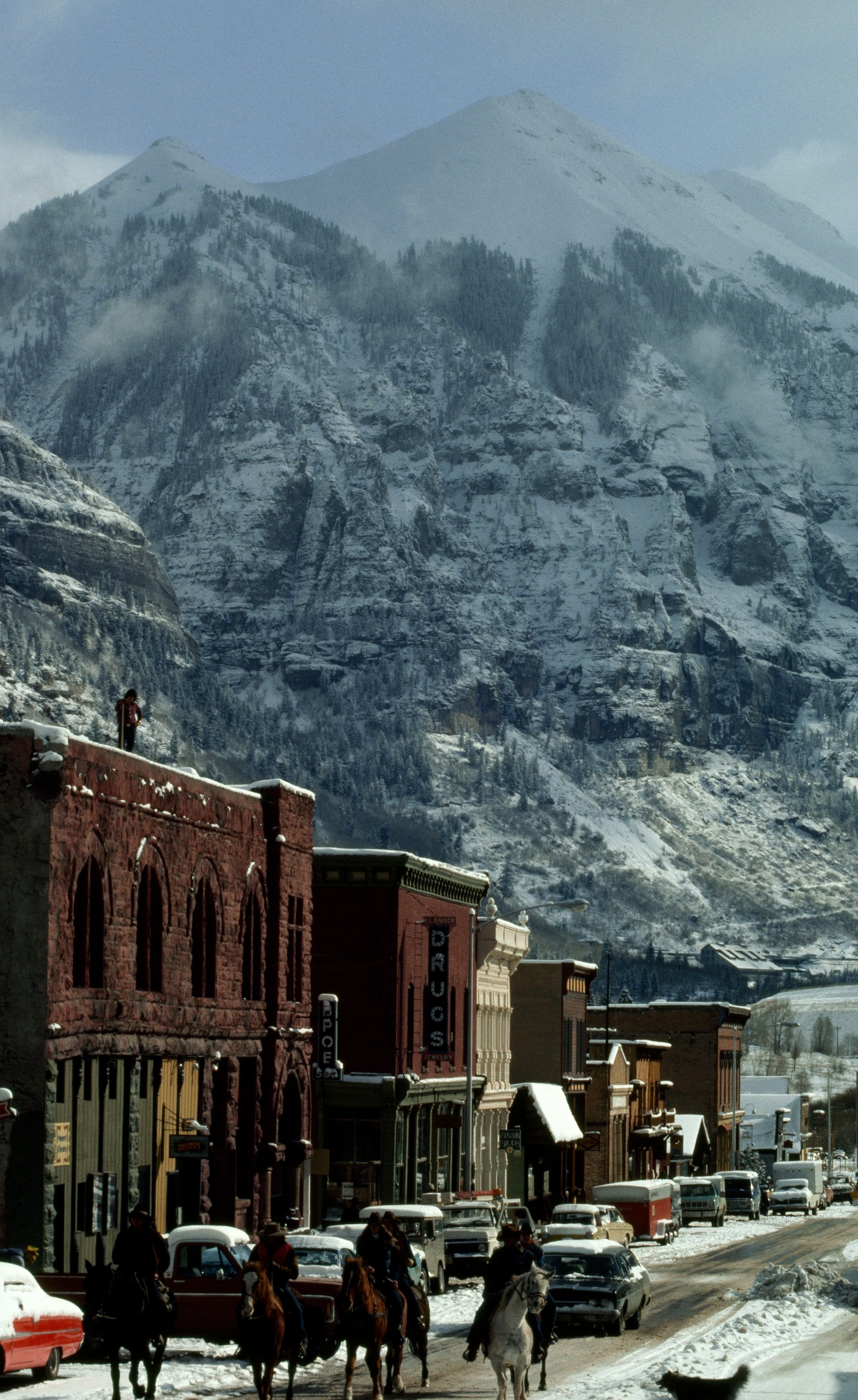 A group of horseback riders walking down the road of a small town with mountains in the background