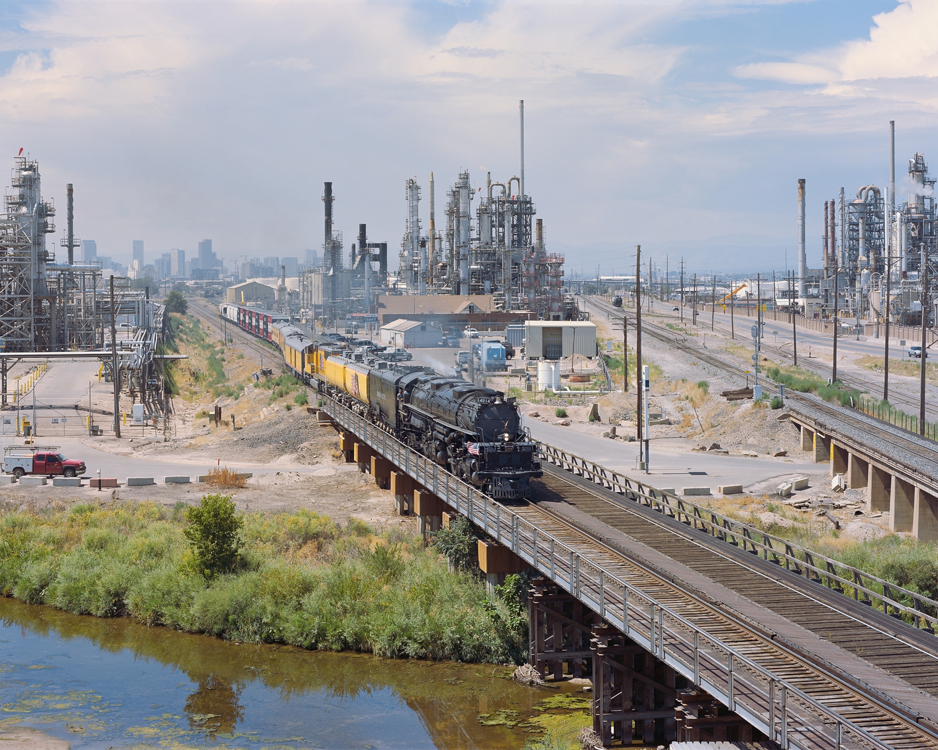 A steam locomotive train moves through an oil refinery, with a city skyline in the background.