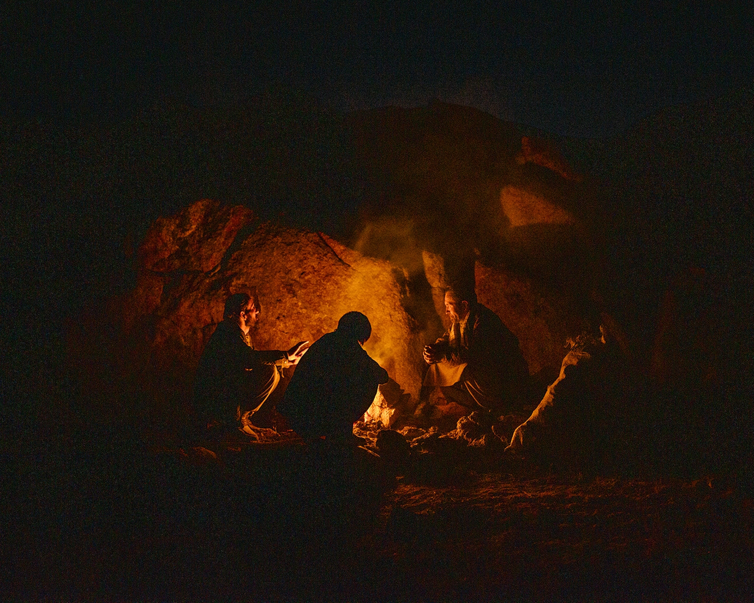 Three people sit around a fire outside at night time as the fire is against a large boulder.