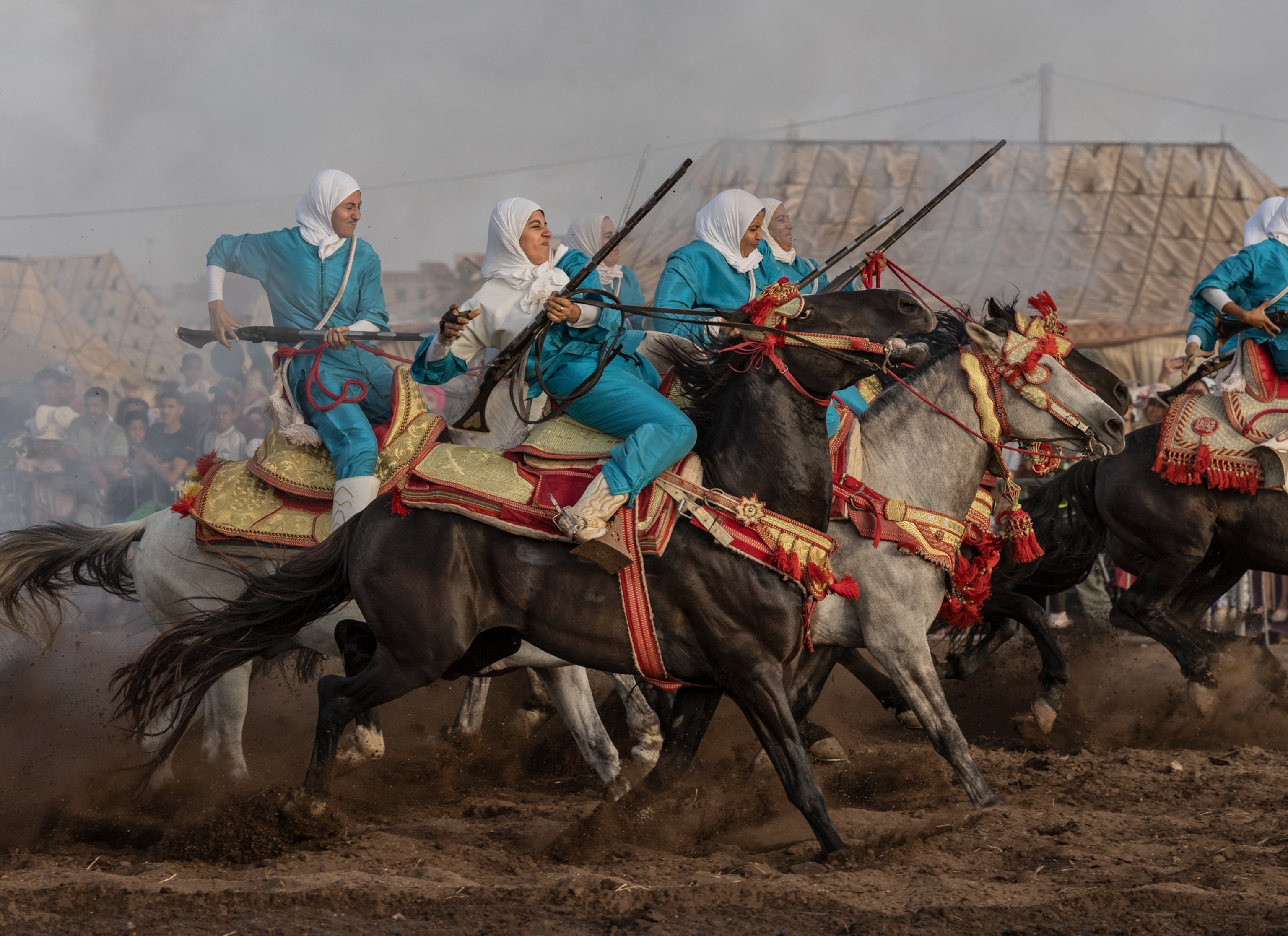Women wearing blue and white head coverings right their horses through dirt. The horses are running and kicking dirt up behind them as the women all have their guns in their hands.