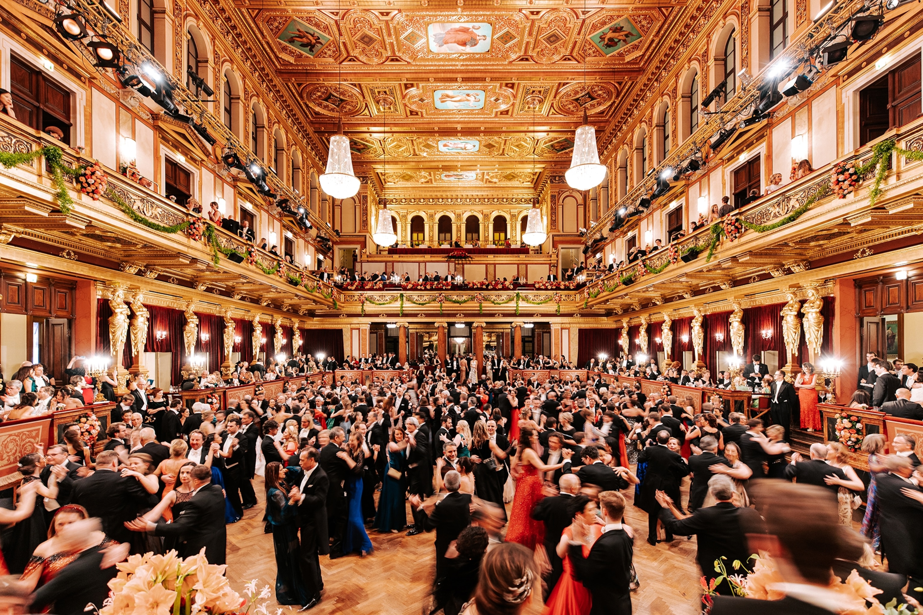 A grand ball room filled with a crowd in evening dresses and suits, dancing in the centre.