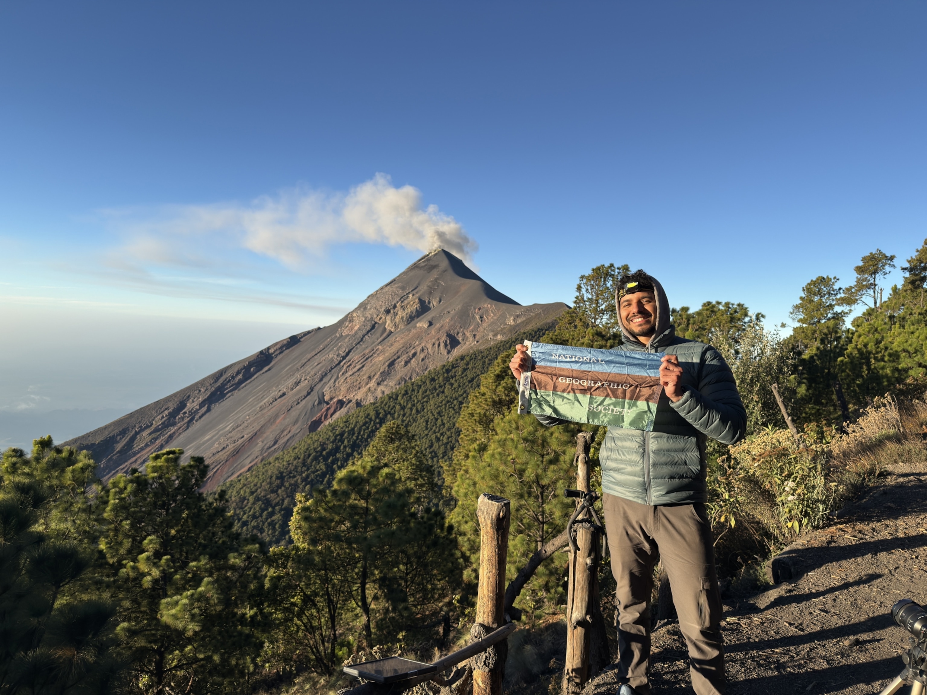 A person smiles while holding a banner at a scenic viewpoint, with a smoking volcano and lush greenery under a clear blue sky in the background.