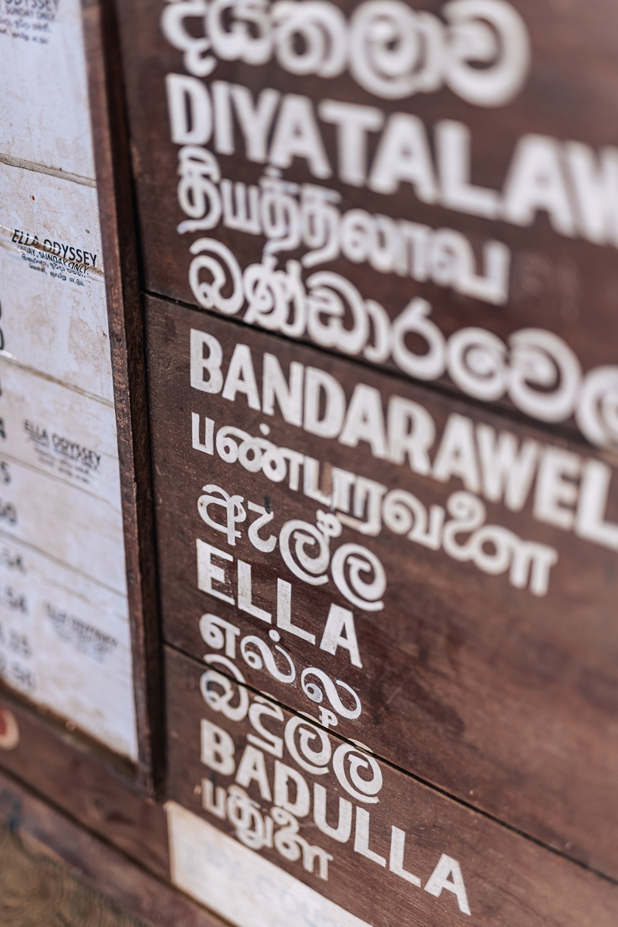 A close-up on a wooden sign at a train station listing multiple stops, including ELLA.