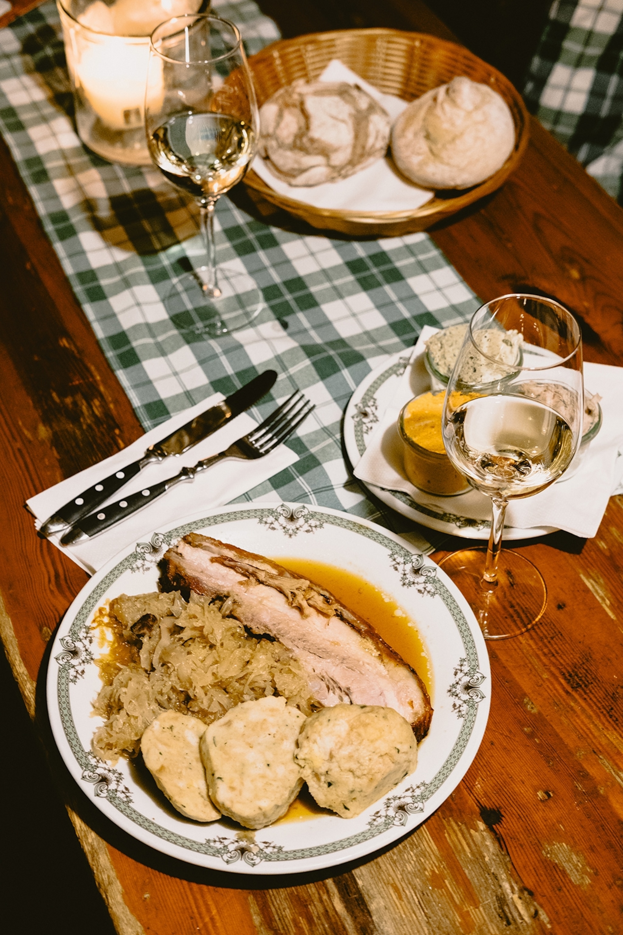 A rustic plate of pork belly with sauerkraut and cut bread dumplings with a side of condiments and wine.