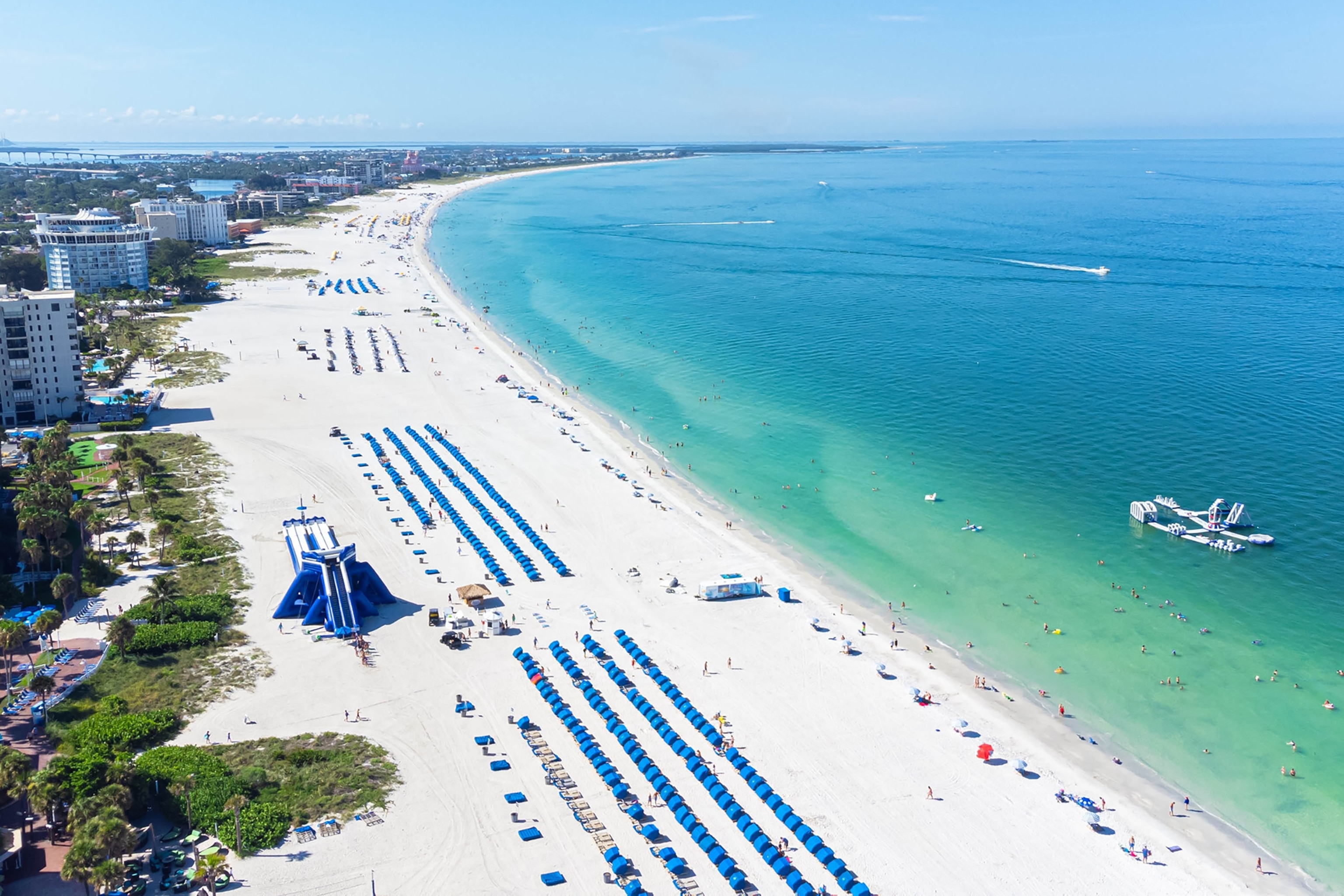 Elevated image of blue umbrellas and sunloungers lined up in blocks along a white sand beach, backed by high-rise buildings and facing a vibrant turquoise sea