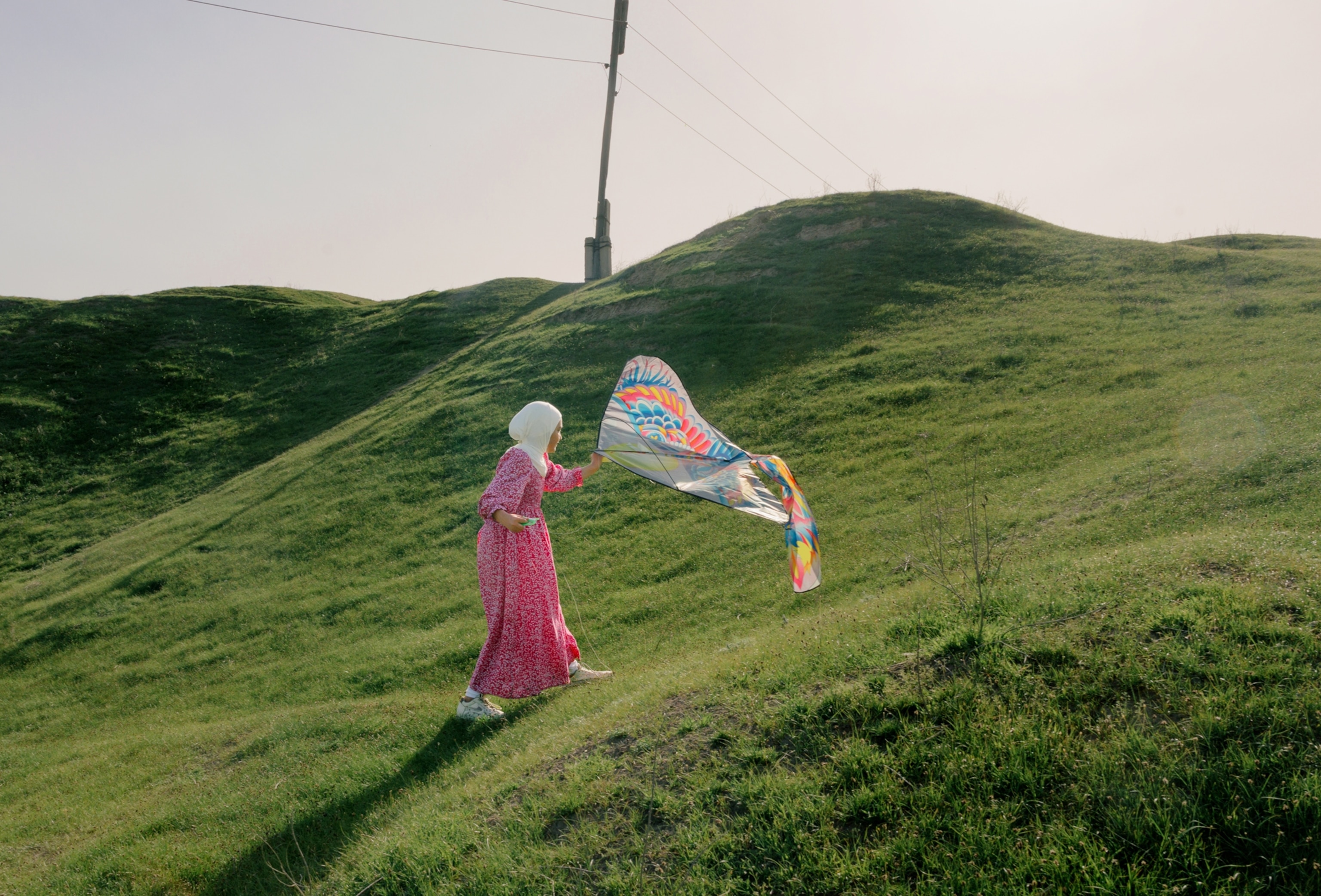 A large, hilly, grassy field with a pink sky in the background and telephone pole with wires. I girl runs in the middle of the field while playing with a kite.