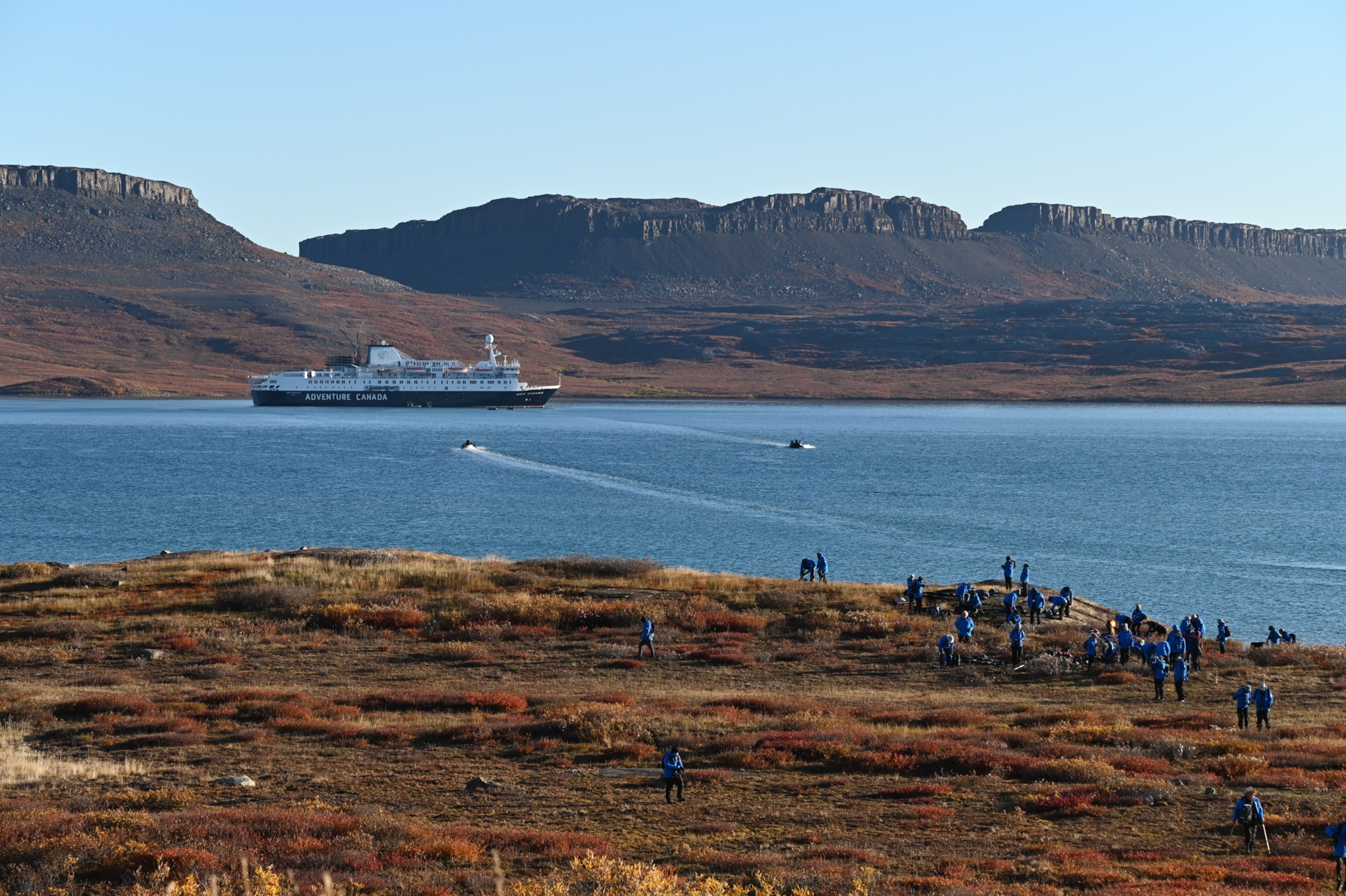 An Adventure Canada cruise ship anchored in a bay near rocky hills, with small boats nearby and a group of people in blue jackets exploring the shore.