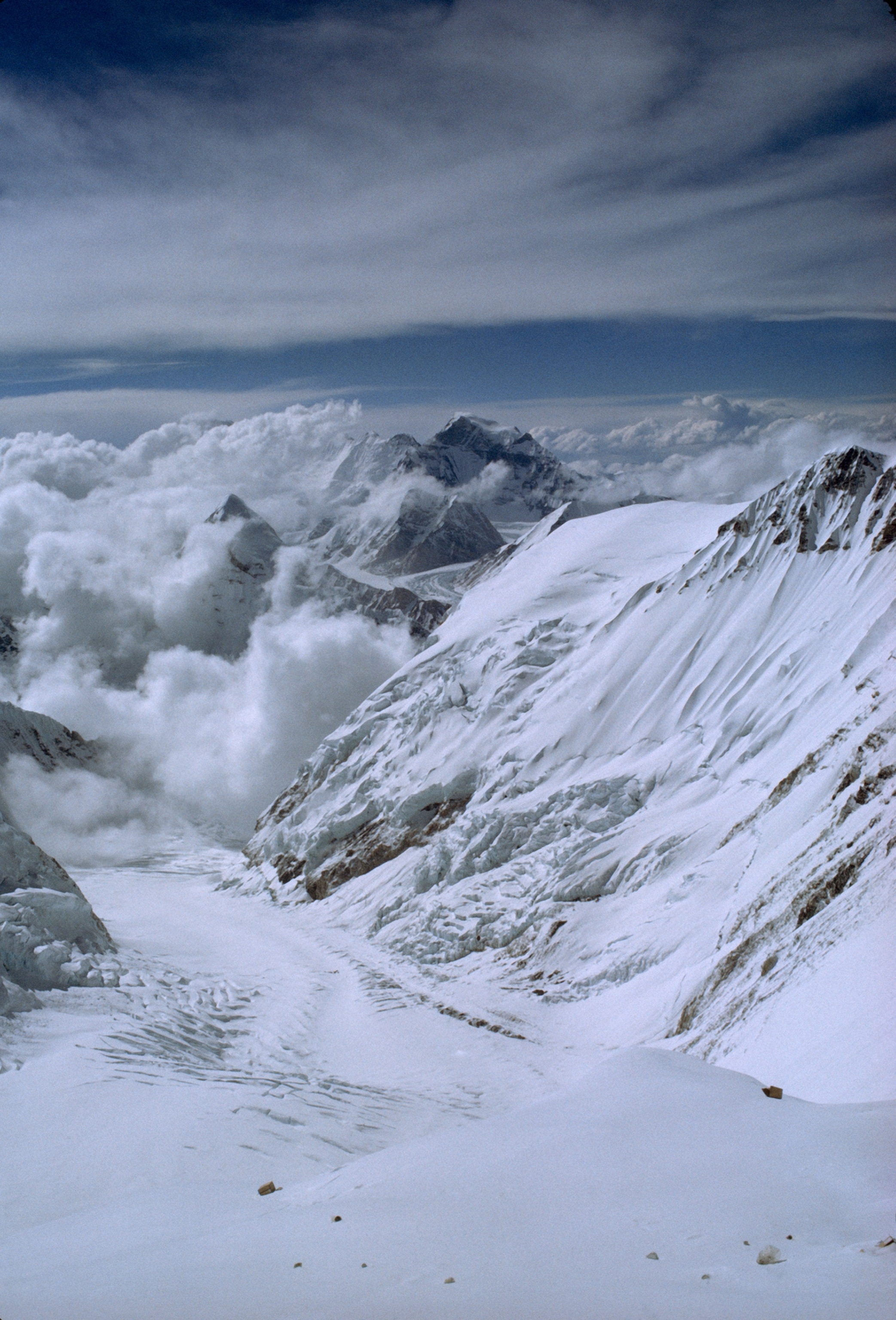 Empty food cartons blow forlornly down the glaciated face of Lhotse. Gazing into the Western Cwmfrom Camp IV, their Advance Base (blue circle), 3,550 feet below, but the ice cliff hid Camp III. They looked down on 23,442-foot Pumori, the pyramid above the clouds. On the right, Everest’s icy west shoulder plunges into the valley.