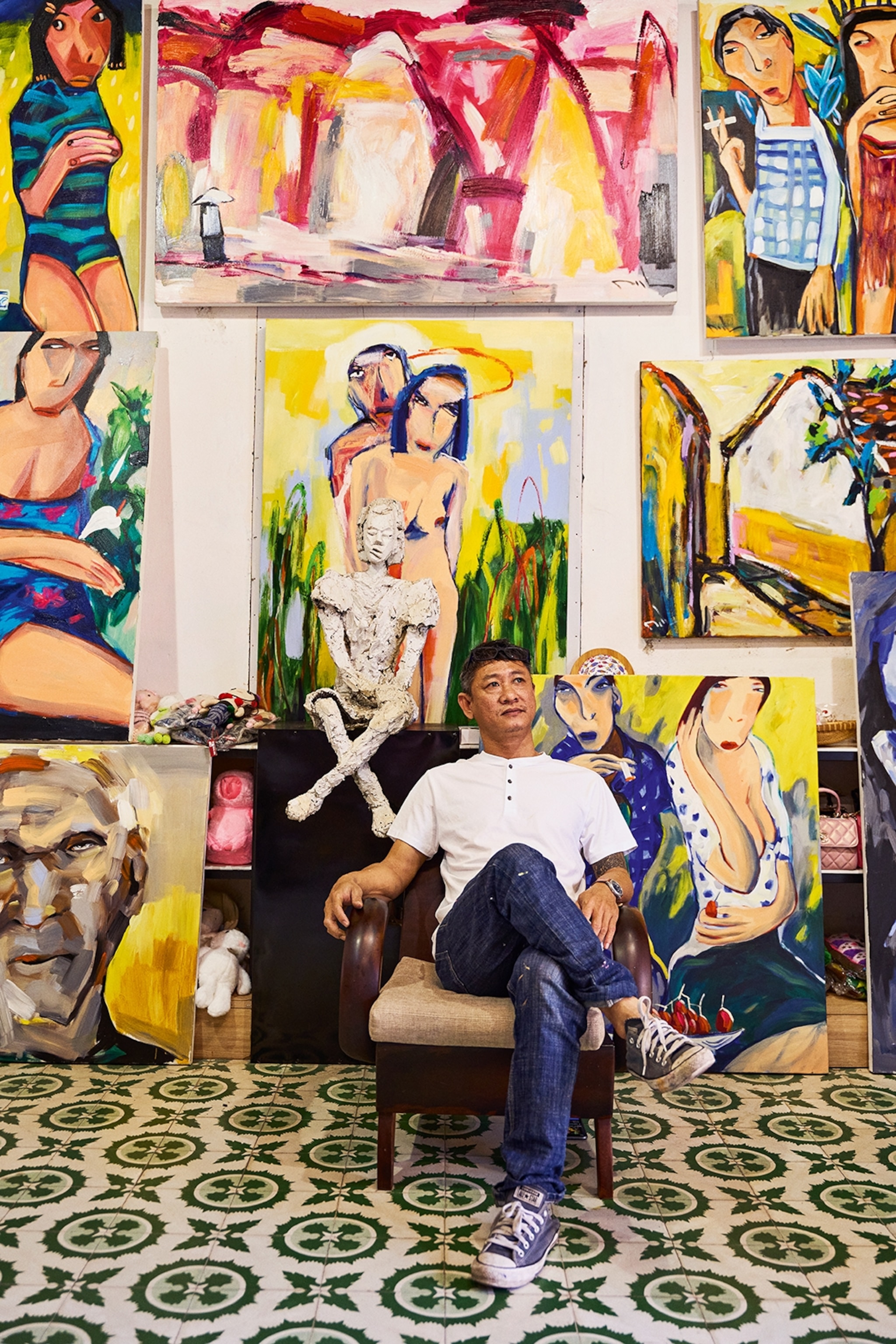 A local painter in simple t-shirt and denim trousers sat on an arm chair in an art gallery with paintings on the wall behind him.