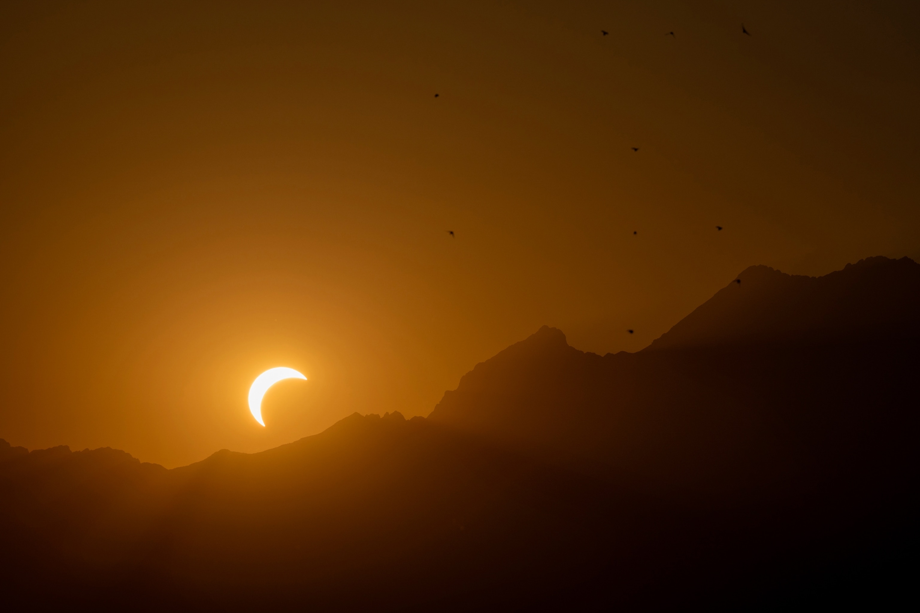 A crescent sun during a partial solar eclipse glows above dark mountain ridges in an amber sky with tiny birds silhouetted overhead.