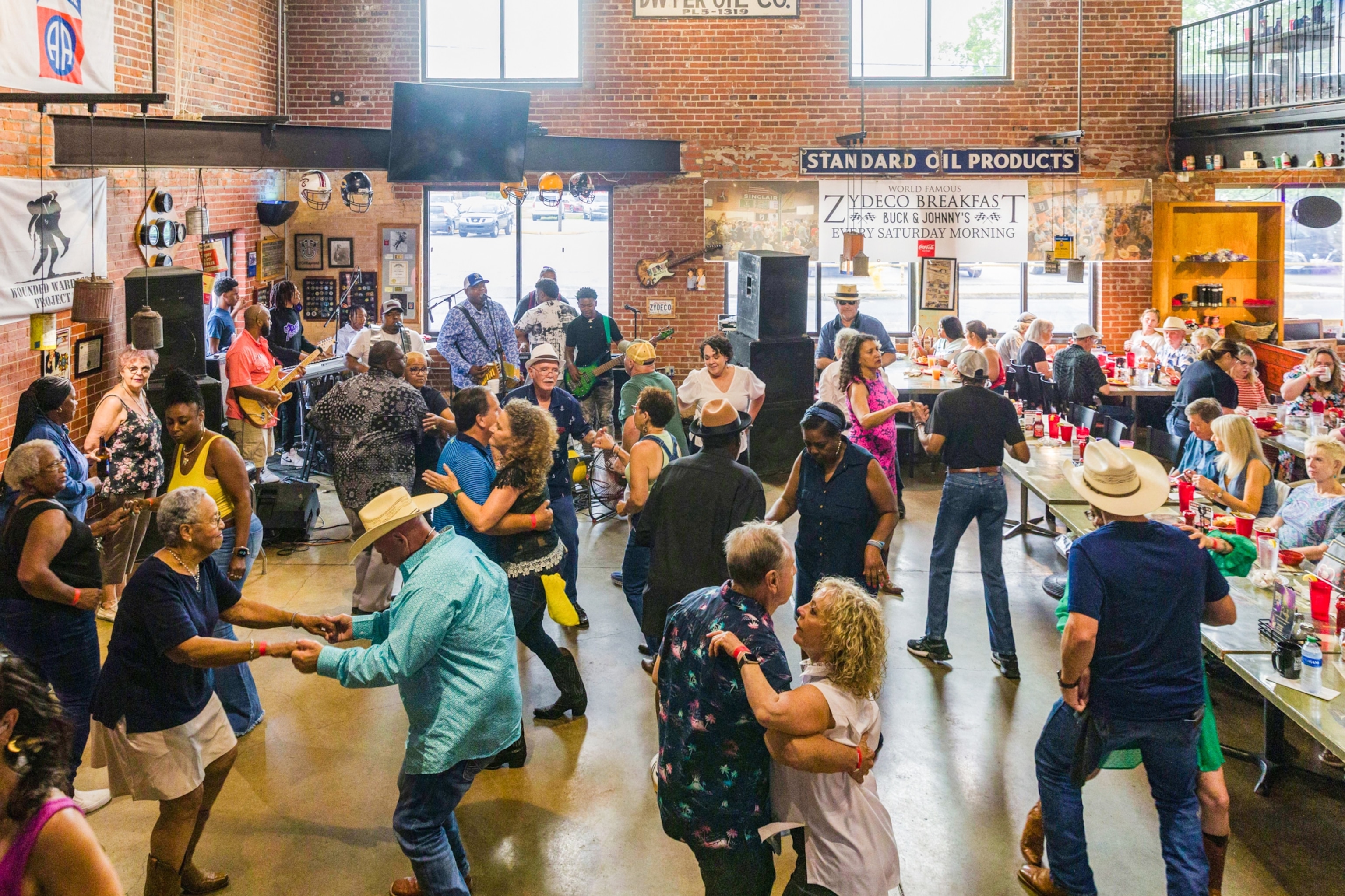 People dance joyfully in a lively bar setting with brick walls. A live band plays near a bar adorned with signs. Patrons seated with drinks.