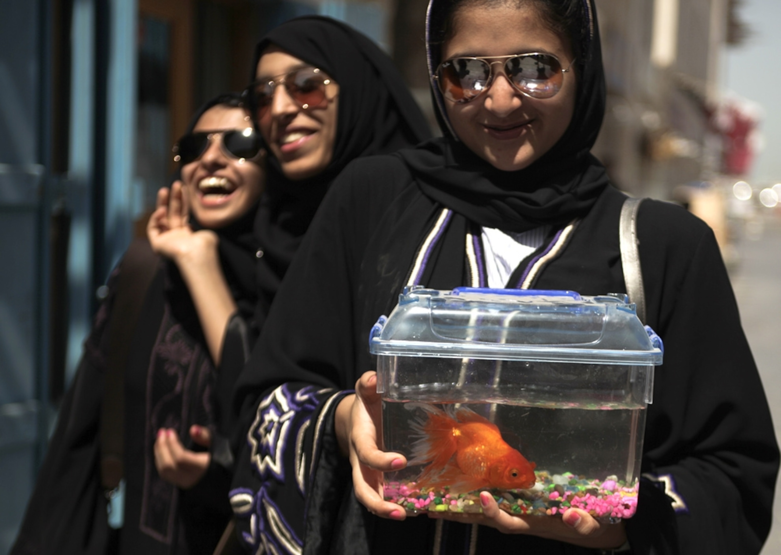 Three Qatari women, wearing abayas, laugh while carrying a pet goldfish.