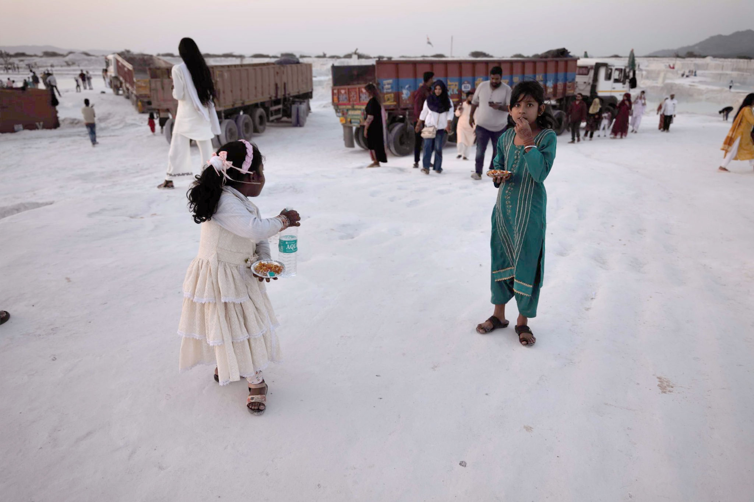 Two young girls eating while on their visit they hold silver plates while standing in front of large trucks.