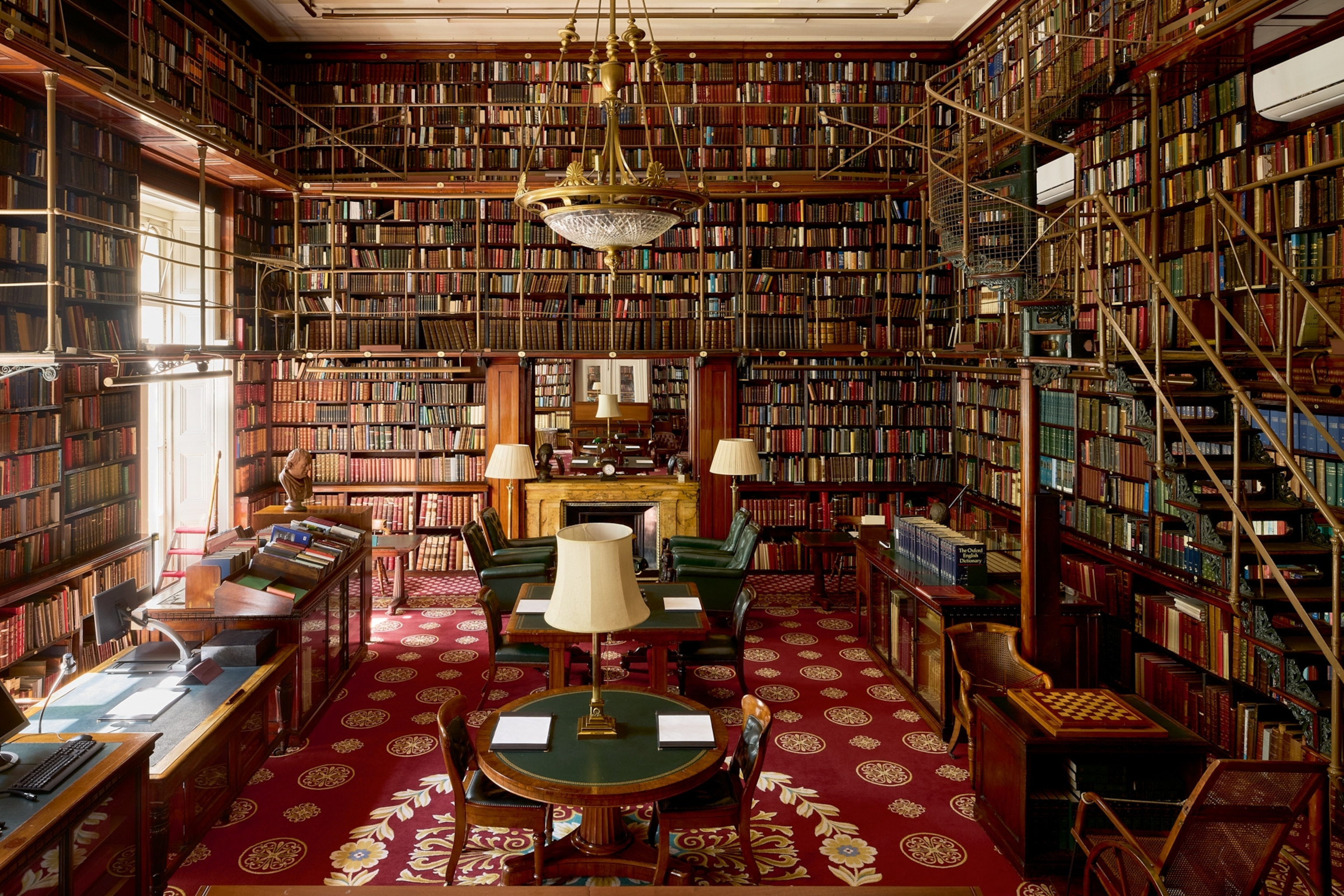 A grand library room filled with floor-to-ceiling bookshelves lined with books. Elegant chandeliers hang from the ceiling, and ornate red carpet covers the floor. Velvet chairs and reading tables with lamps create a scholarly and serene atmosphere.
