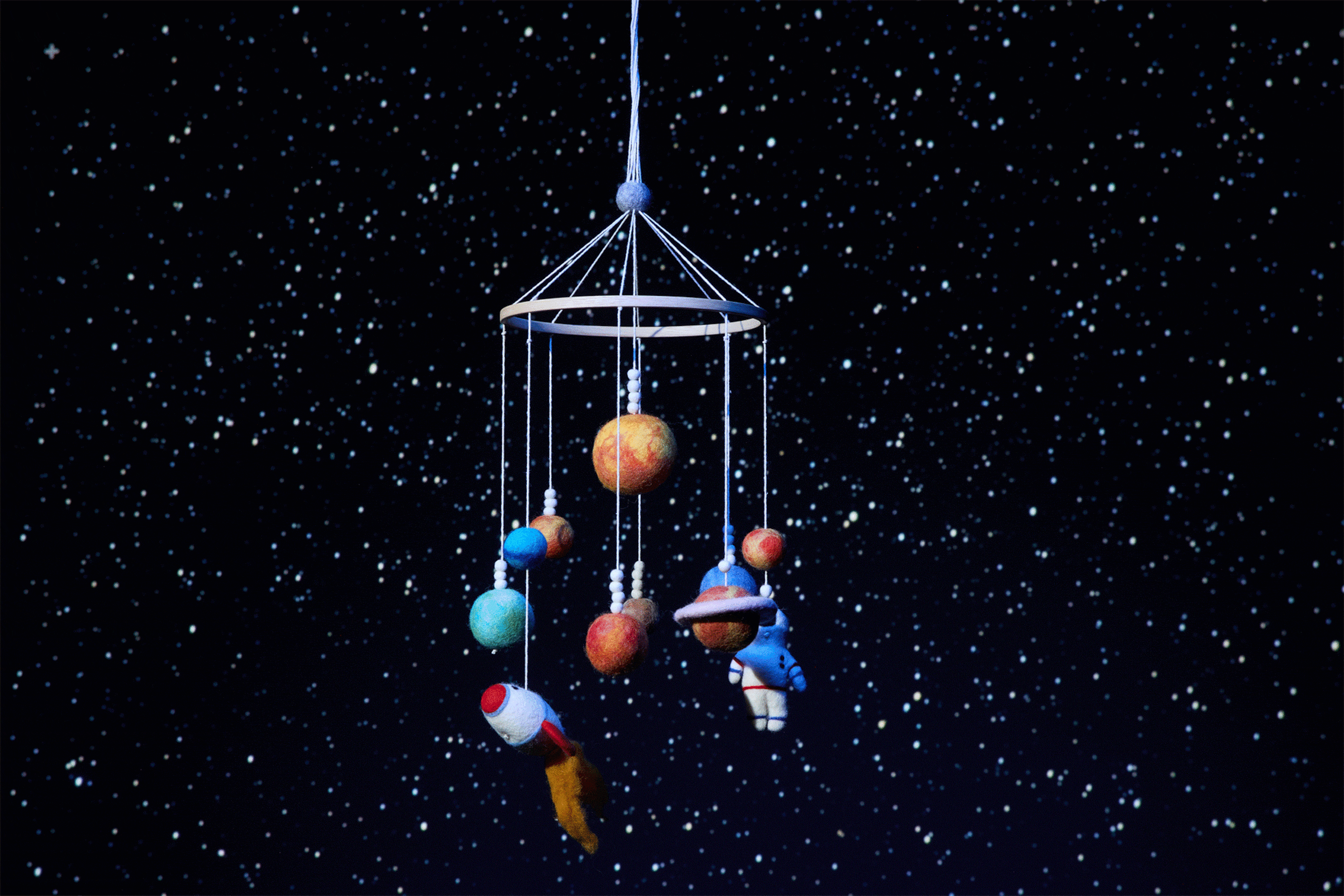 A space-themed mobile with an astronaut, rocket ship, and planets made of felted wool spins in front of a starry sky.