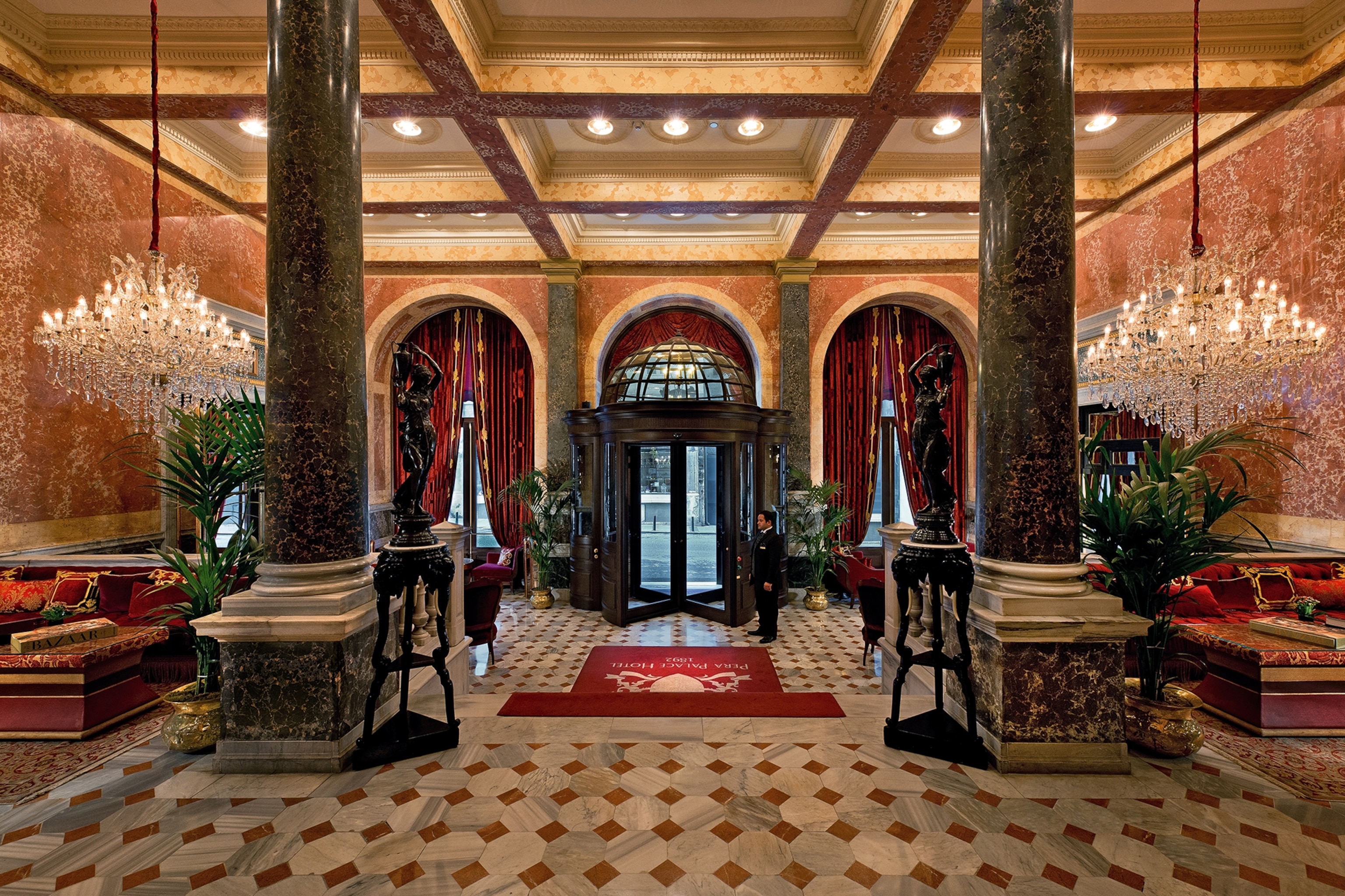 A romantic and luxurious marble-floored entrance hall of a hotel with bronze statues and a red carpet leading the way inside.