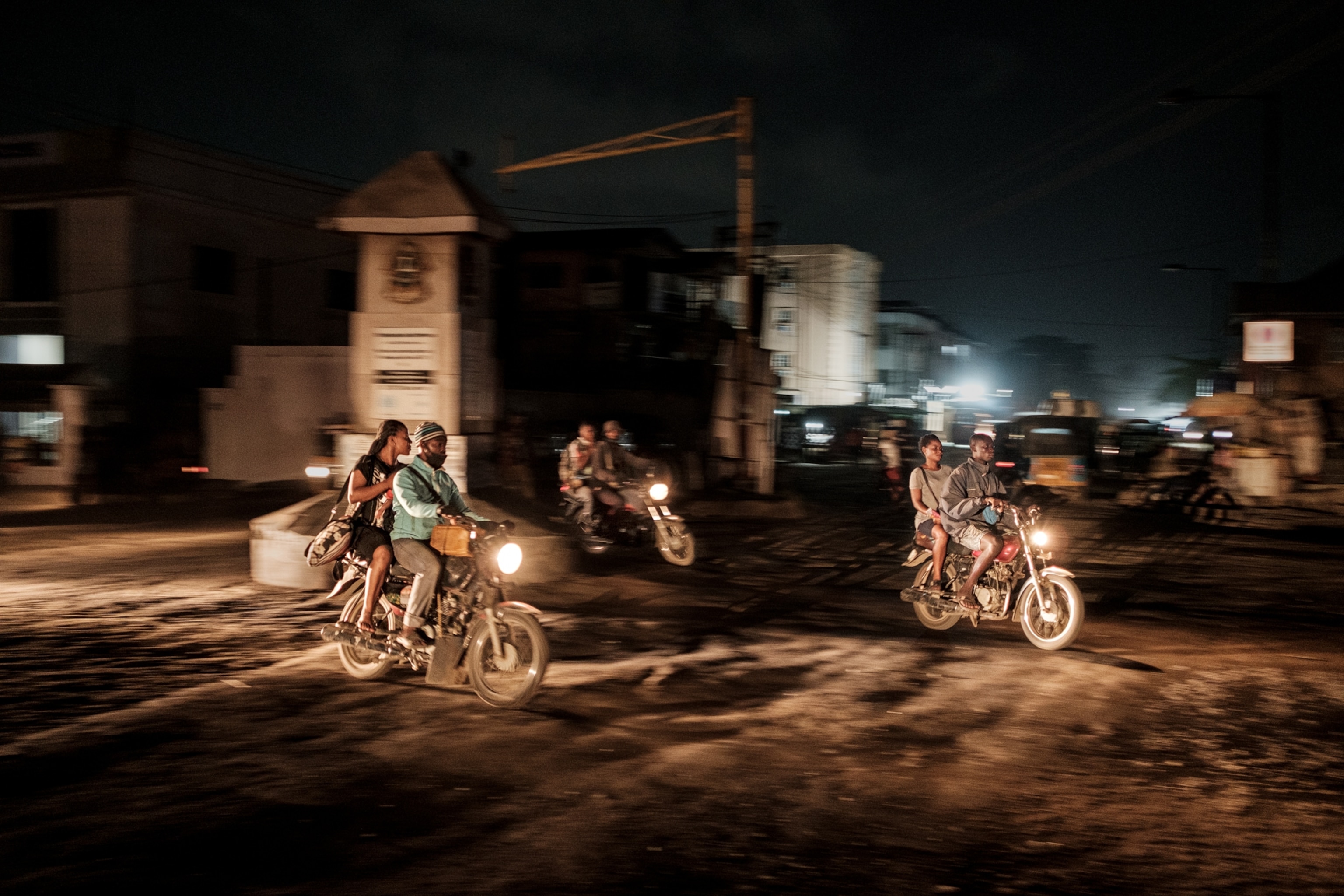 Several motorcycles with passengers on dirt town street at night.