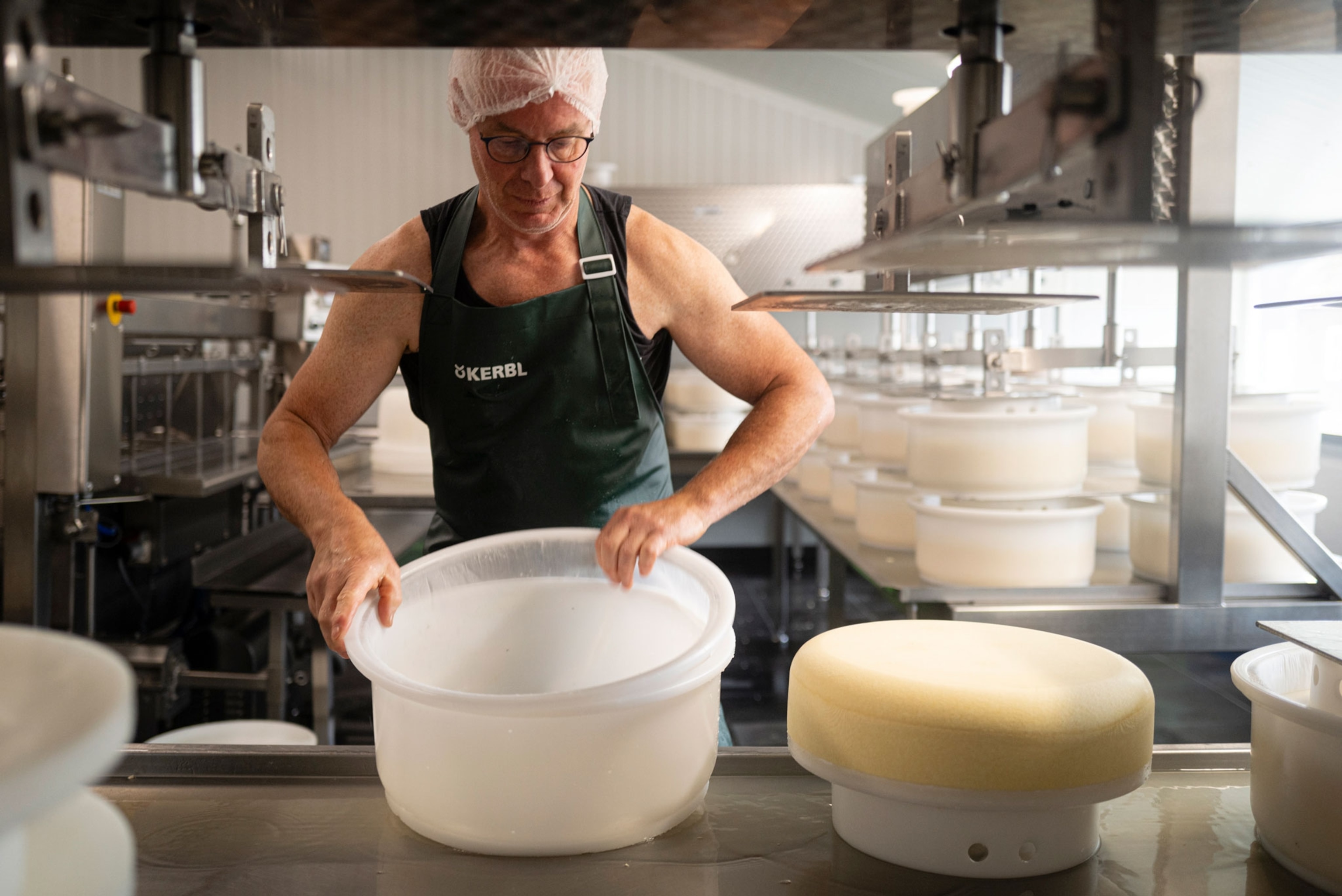 A man wearing an apron and hairnet works in a cheese factory, holding large white molds next to a large light yellow block of cheese on a steel counter.
