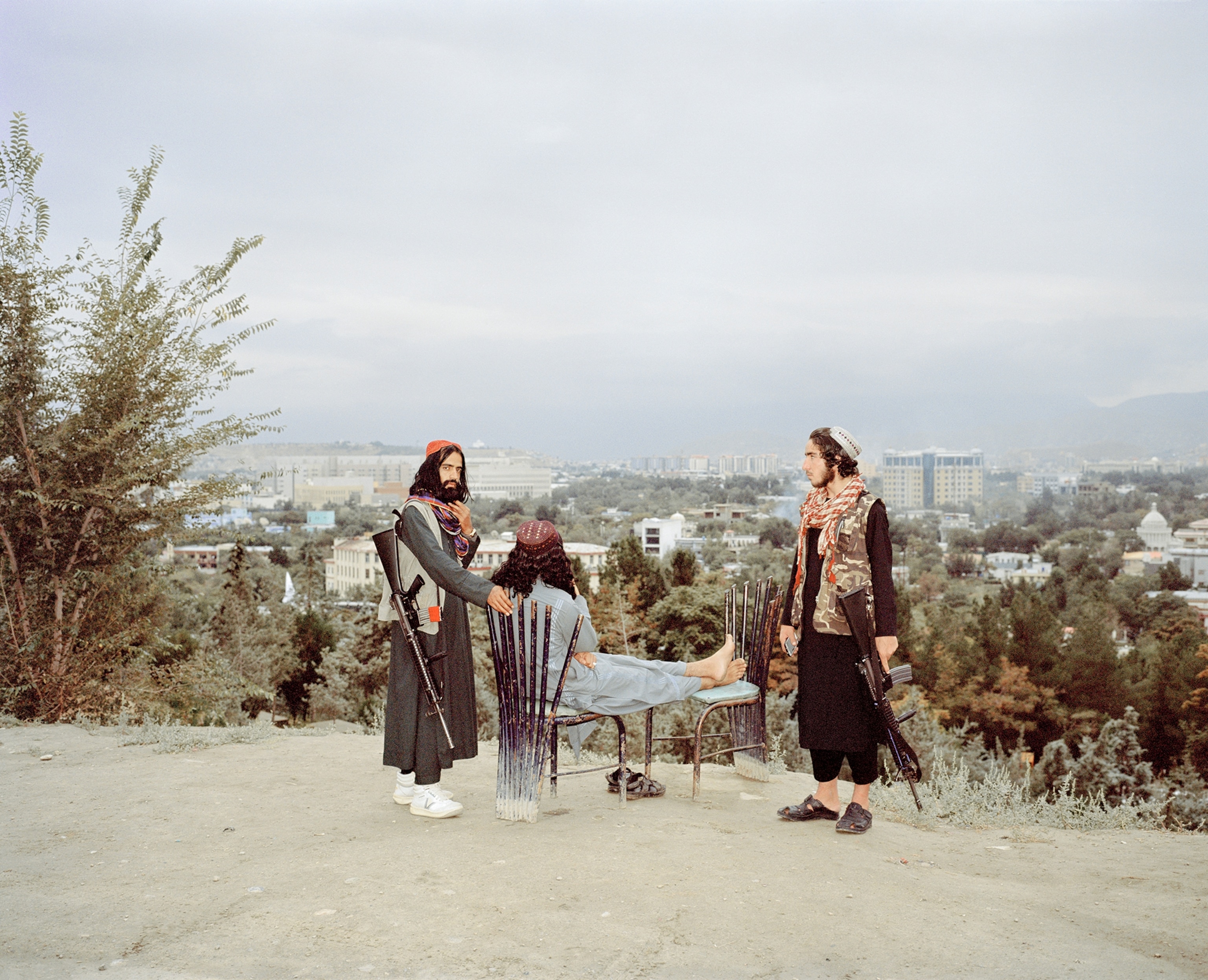 Two young men with guns stand beside a seated young man with the city of Kabul in the background.