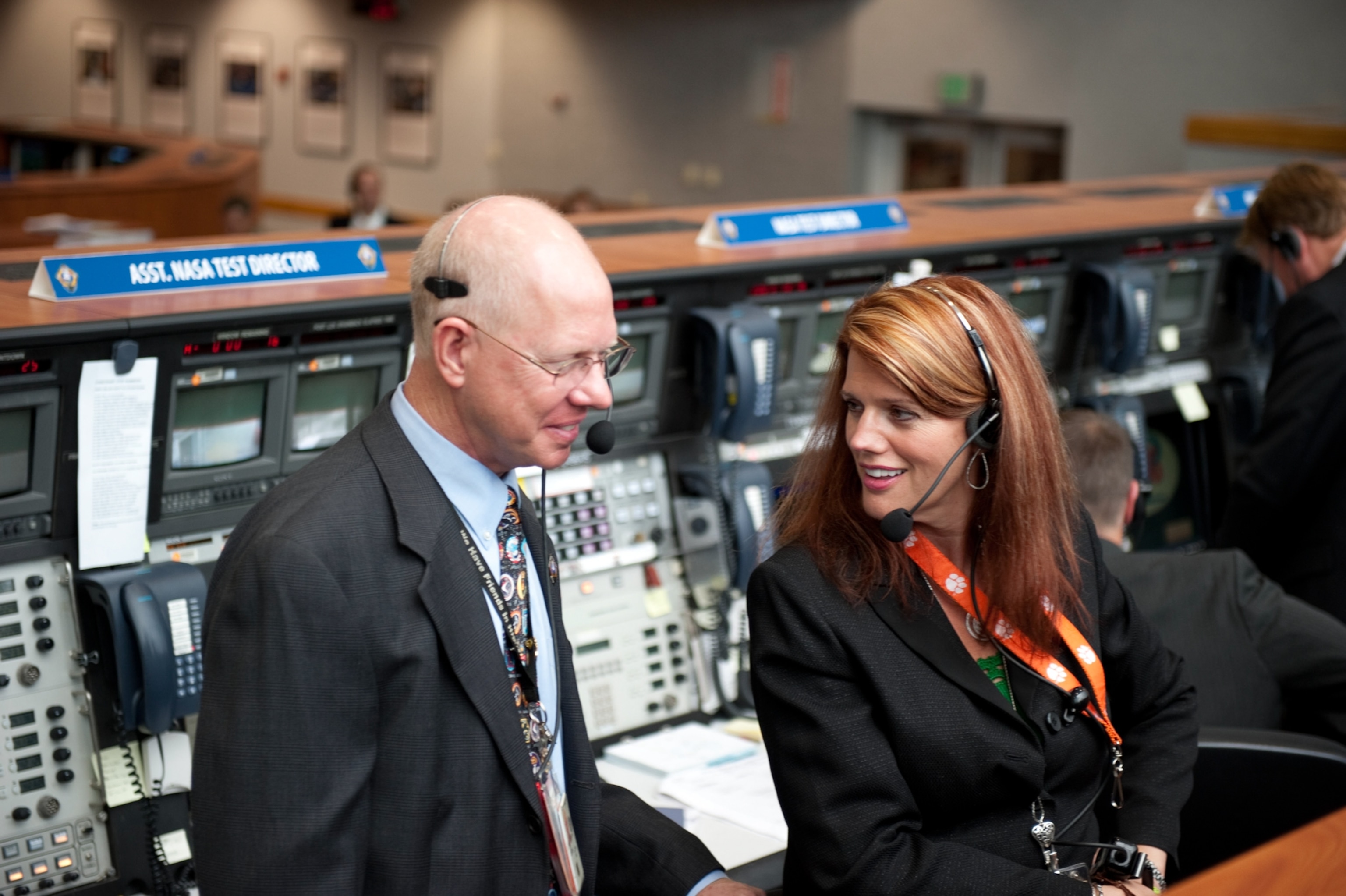 Two people in a NASA control room, both wearing headsets and formal attire, smile while conversing. Multiple monitors and equipment surround them.