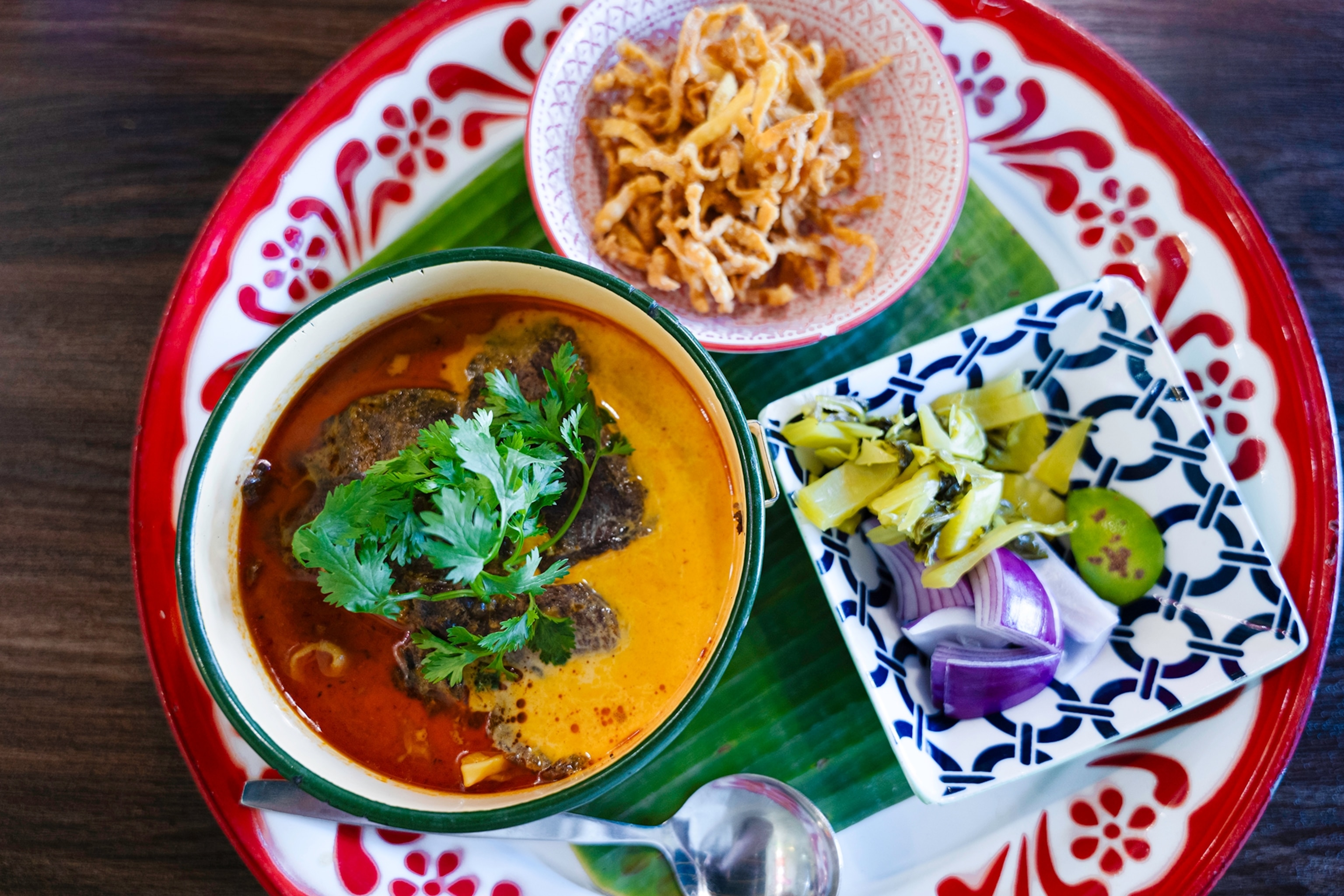 An array of Thai dishes on a decorative plate, including a beef curry with noodles and fresh condiments to add on the side.