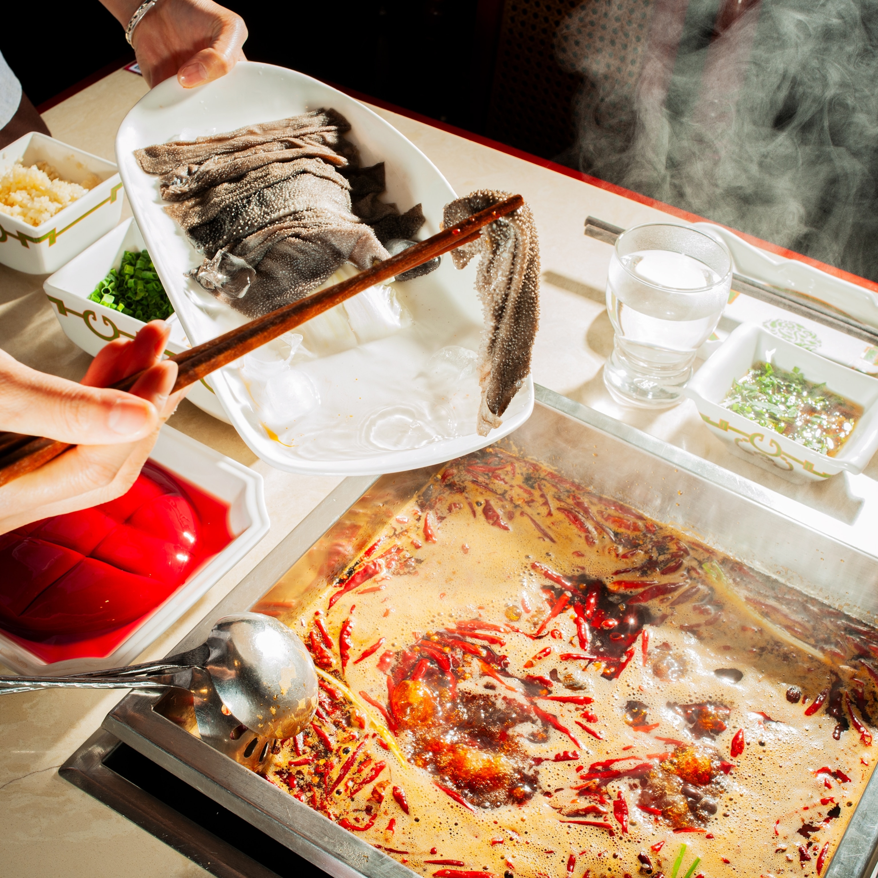 A person uses chopsticks to place thin slices of fish into a steaming hot pot filled with broth, dried chilies, and spices, while small bowls of ingredients sit nearby on the table.