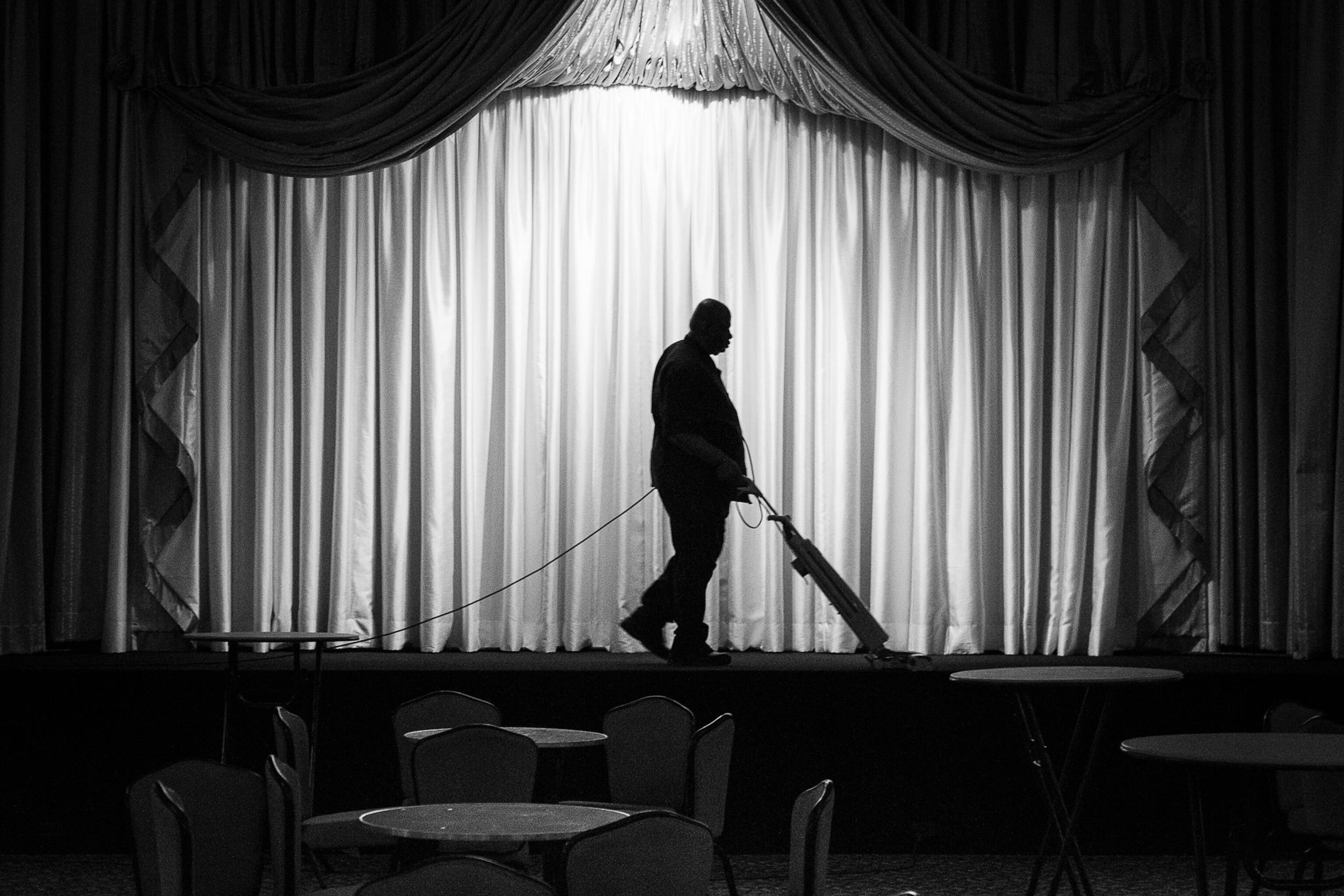 a person vacuuming the stage in a theater, their figure silhouetted against the lighter curtains