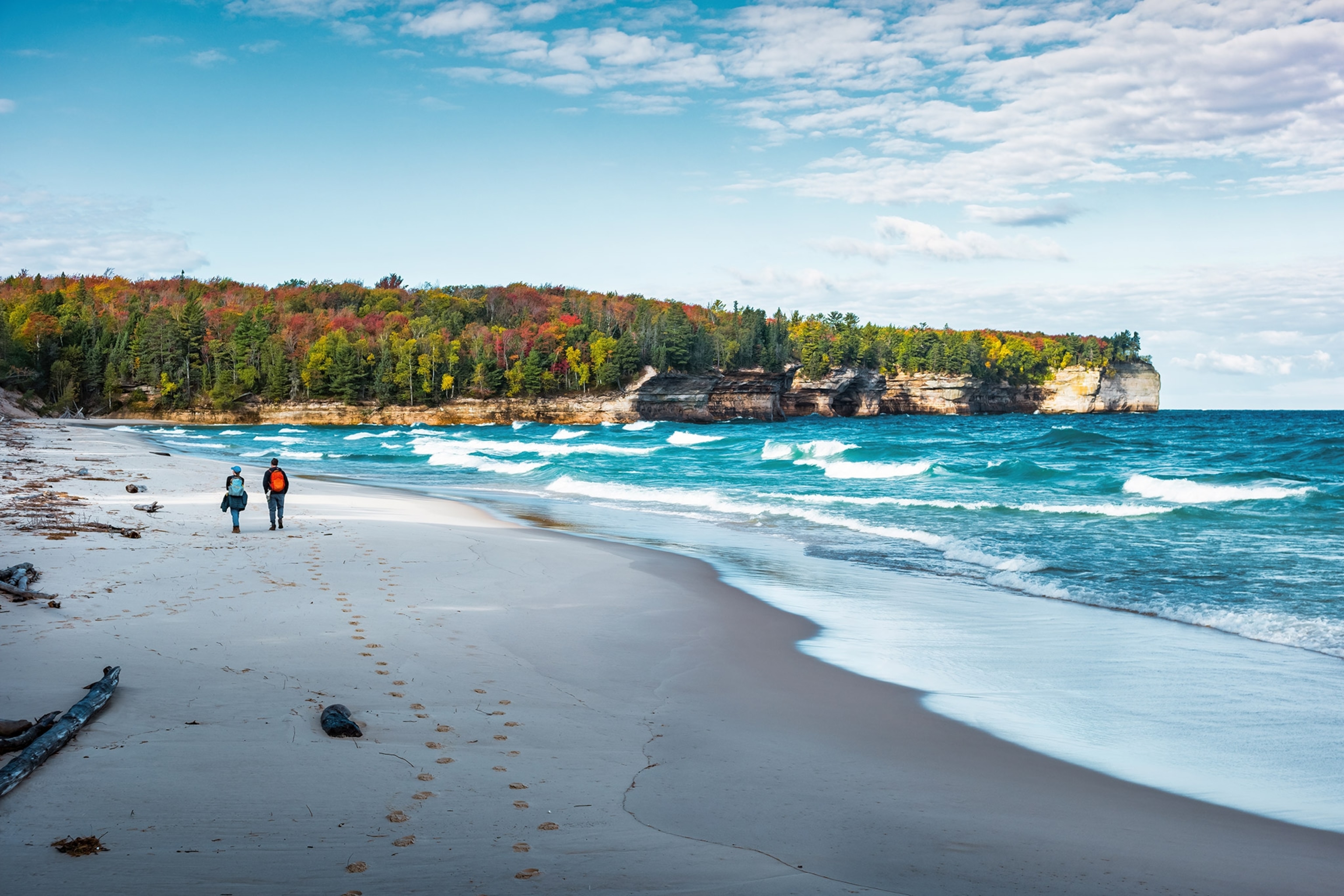 Two people walk along a sandy beach with turquoise waves lapping, wooded cliffs in the distance