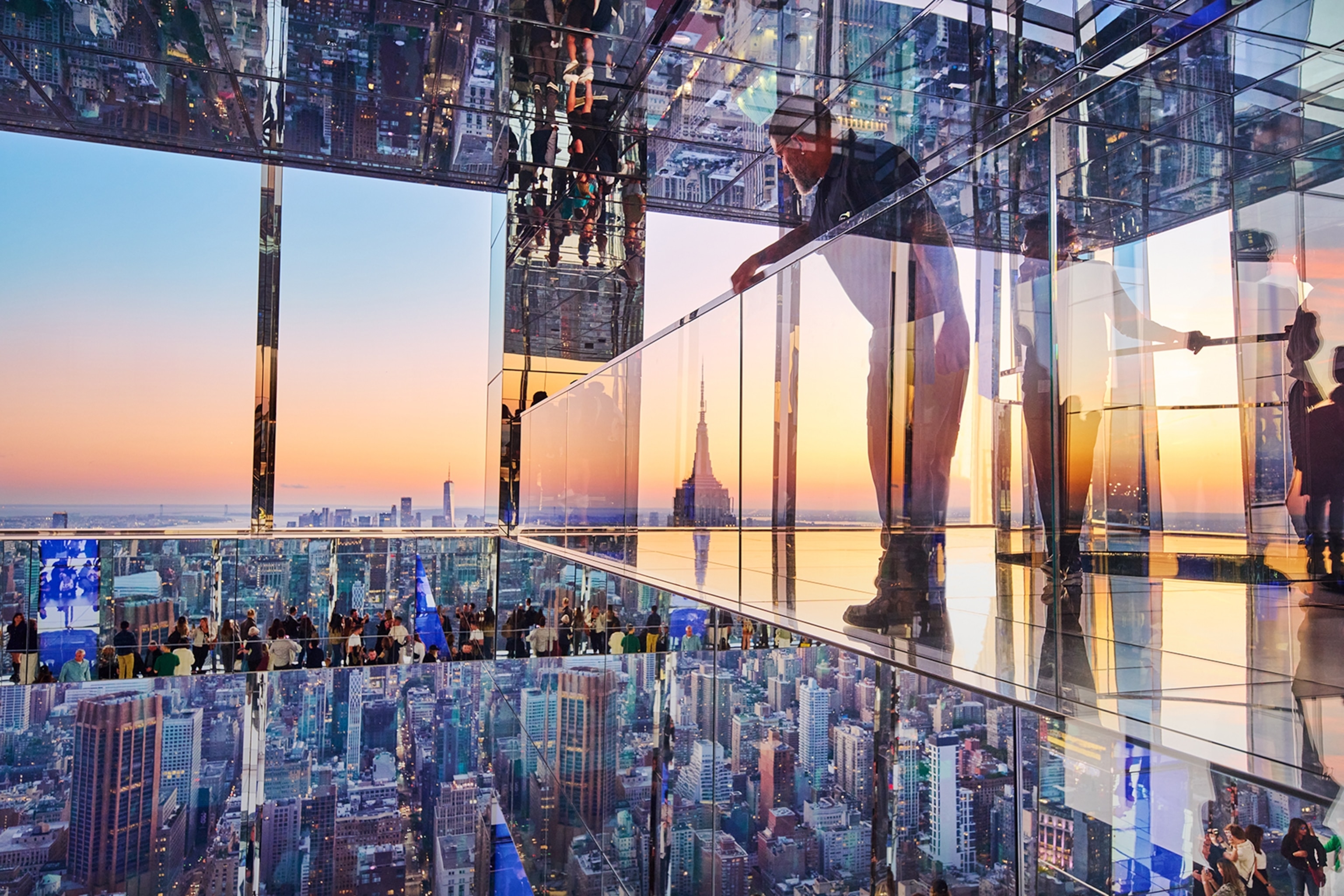 Man standing on a glass platform in a glass building at sunset looking out over a city skyline filled with high-rise buildings