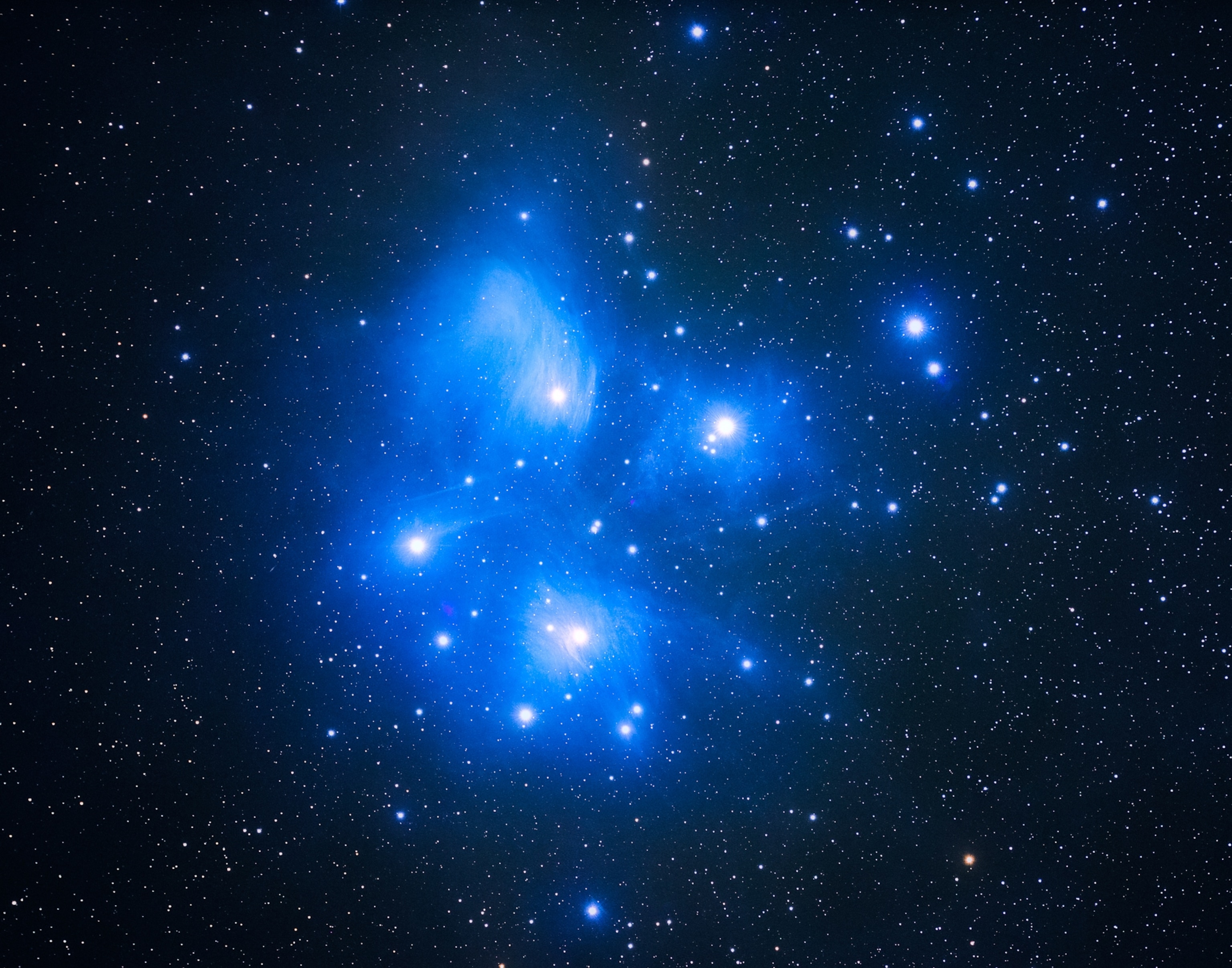 A bright blue star cluster shines against a dark sky, with hundreds of stars surrounded by glowing clouds of blue dust.