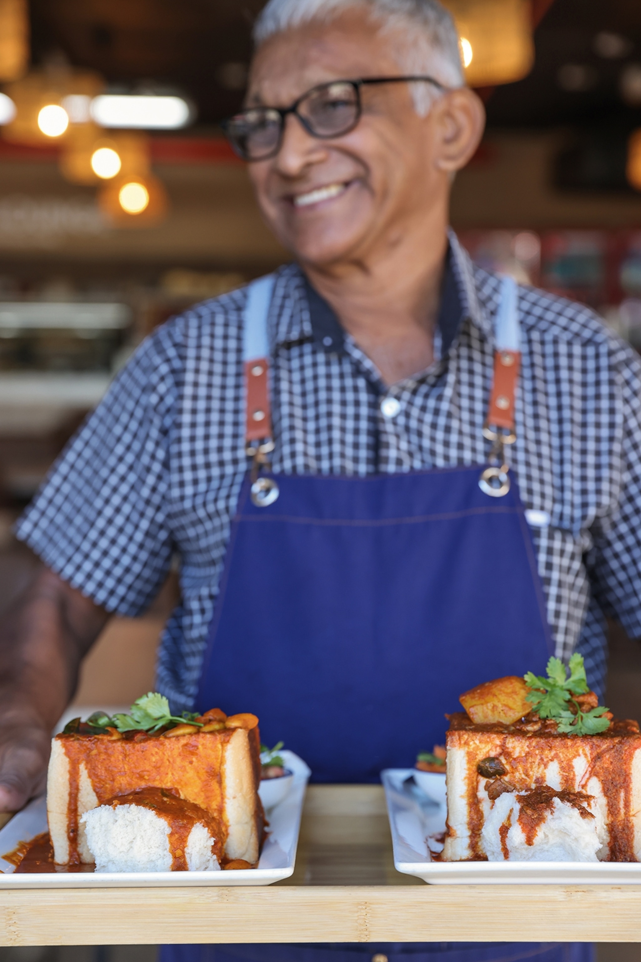 A smiling man wearing a canvas apron serving a tray of two bunny chow curry buns.