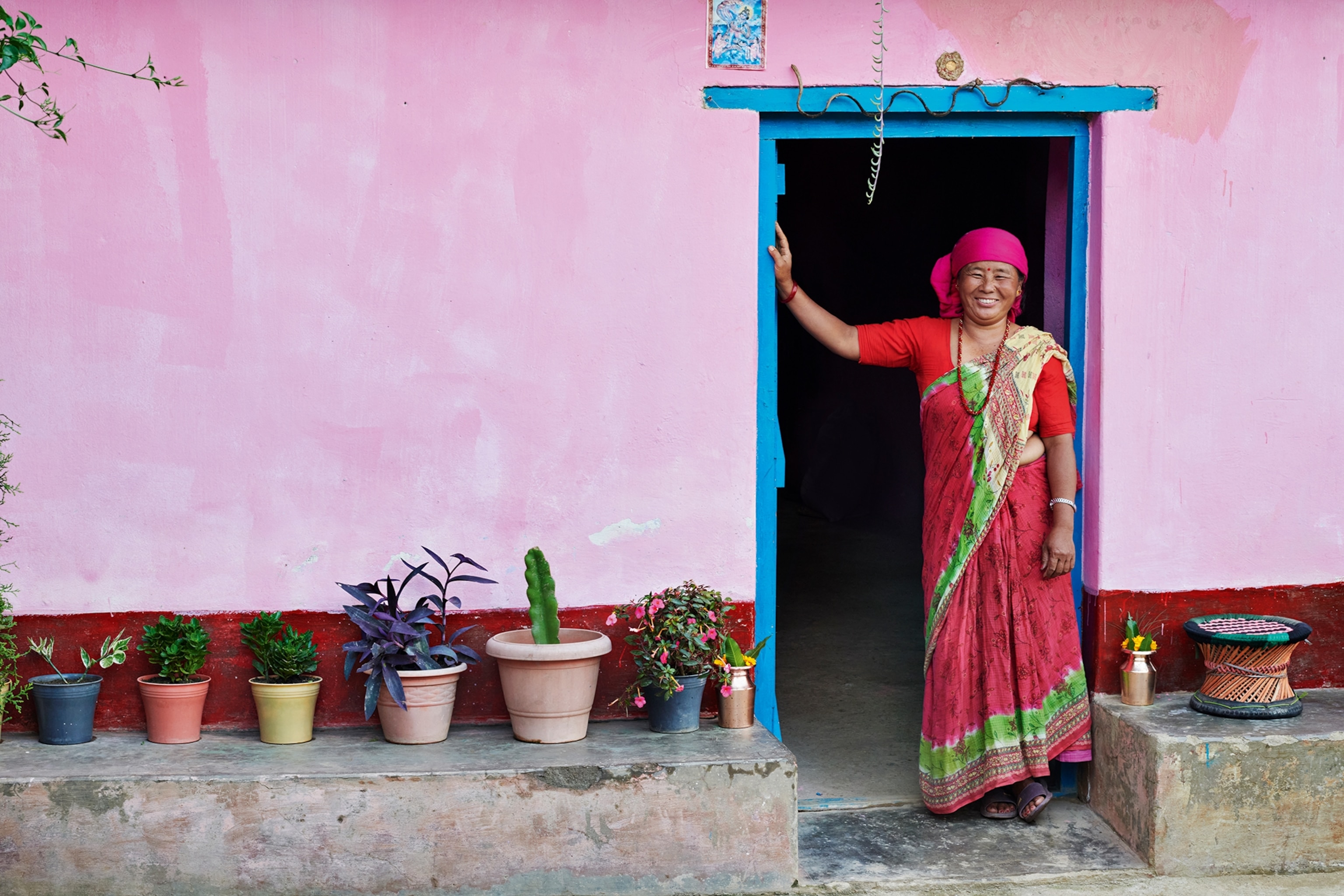 A friendly, smiling woman leaning with her stretched-out arm against the door frame of her house, wearing a traditional Nepalese sari and colourful headwrap.