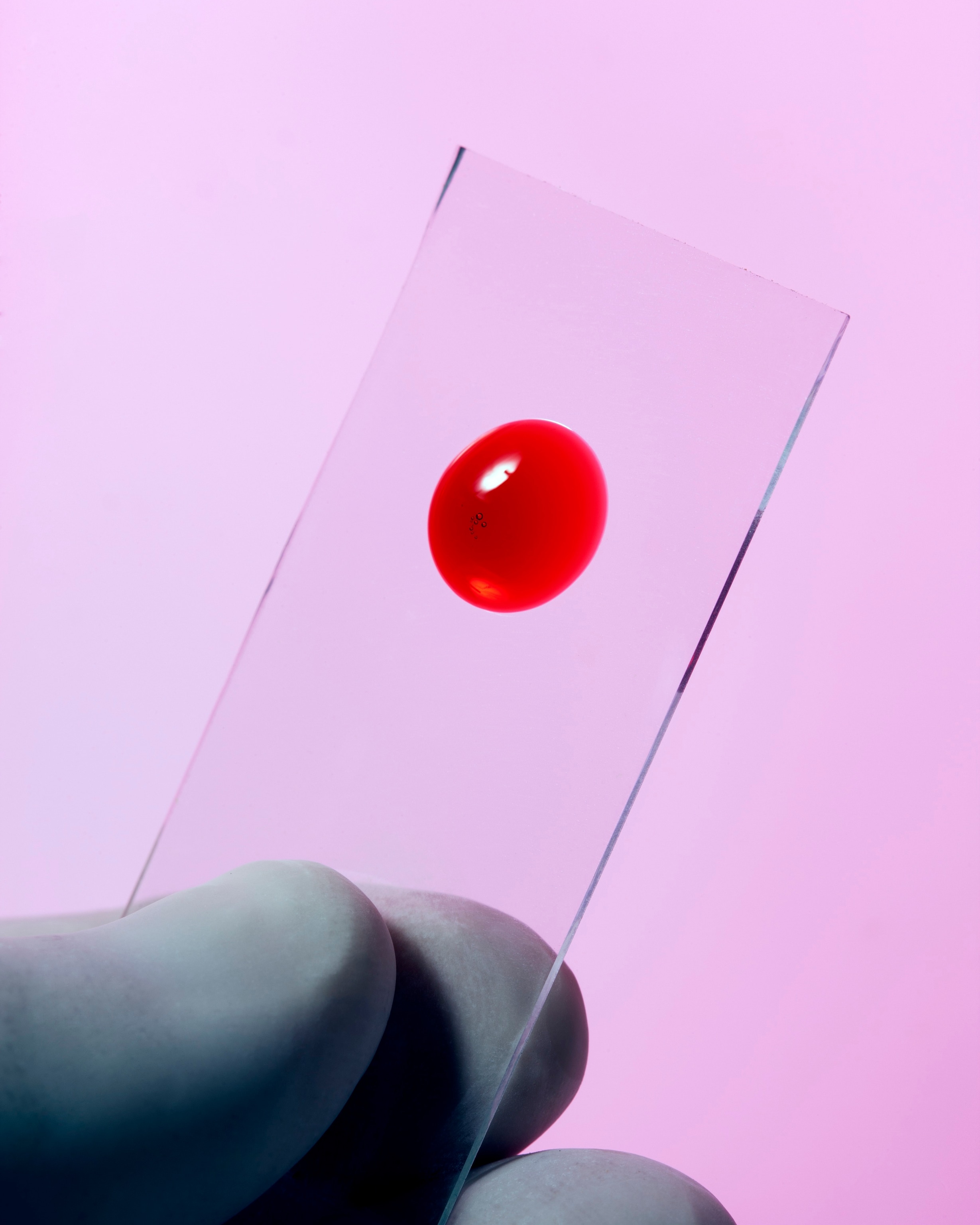 A gloved hand holds a rectangle glass slide with a drop of blood in the center against a pink background