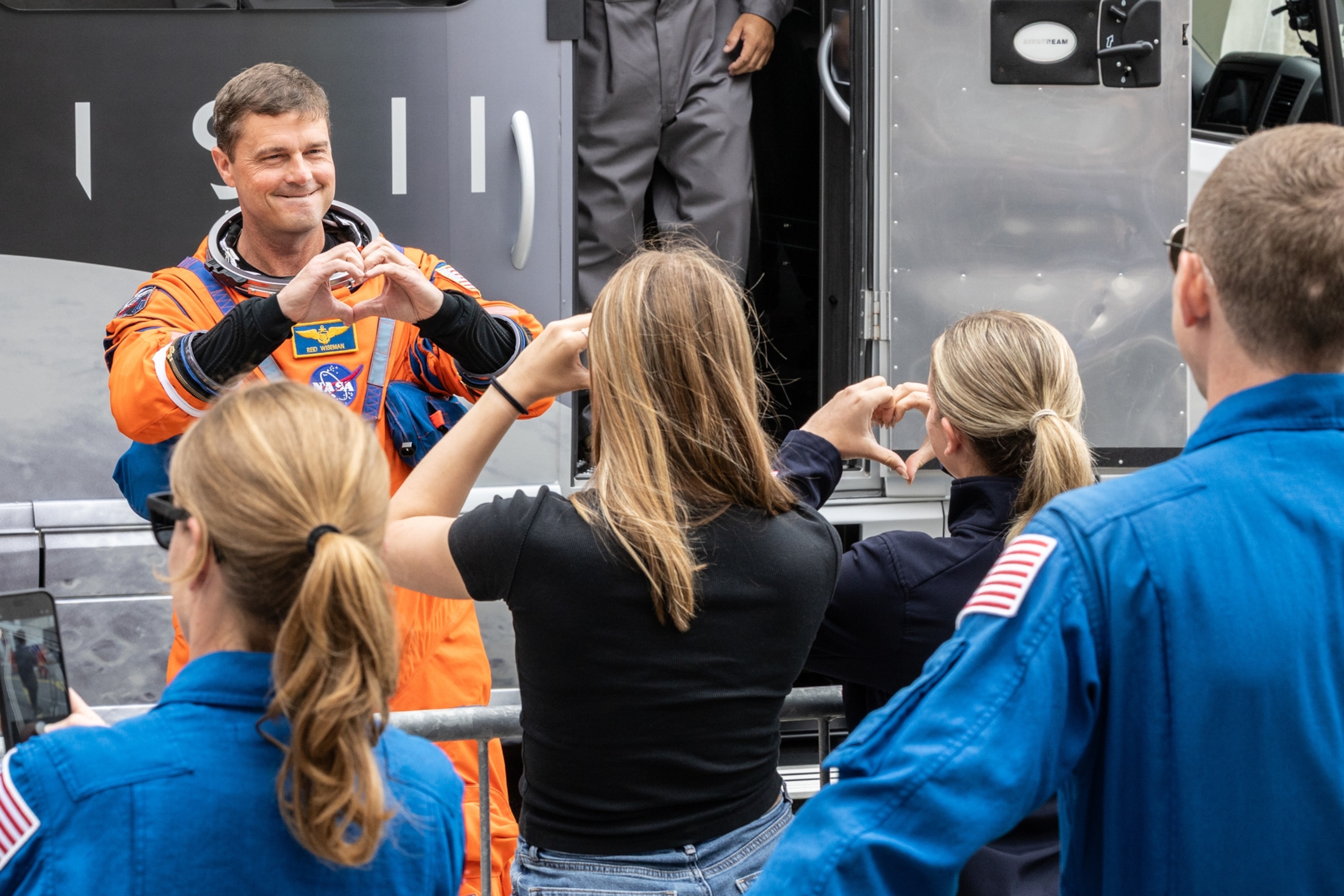 Astronaut in an orange suit makes a heart gesture, smiling at a group, including people in blue suits with U.S. flags