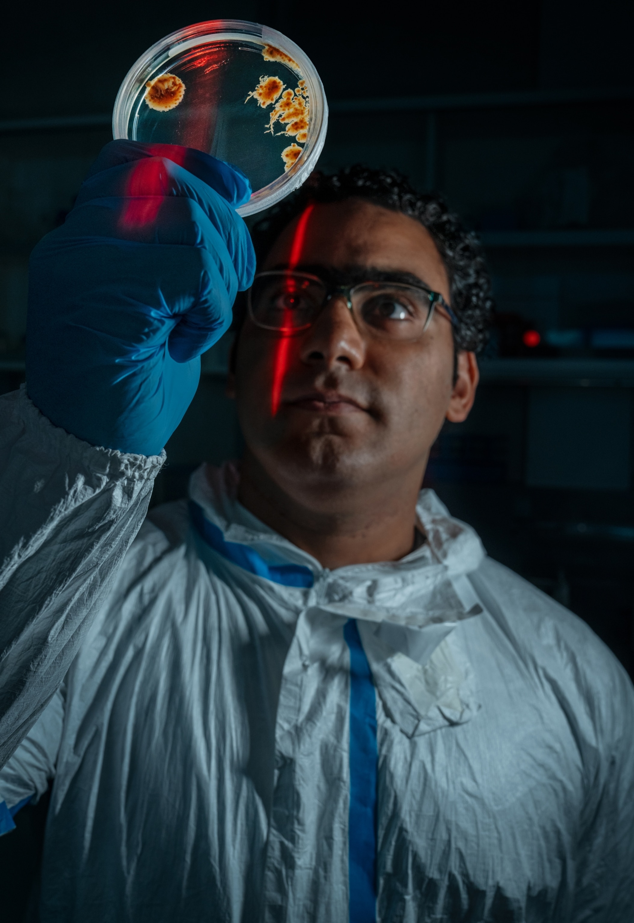 A scientist holds a Petri dish with samples in it in front of his face in a portrait. The scientist is wearing blue gloves and a plastic coat.