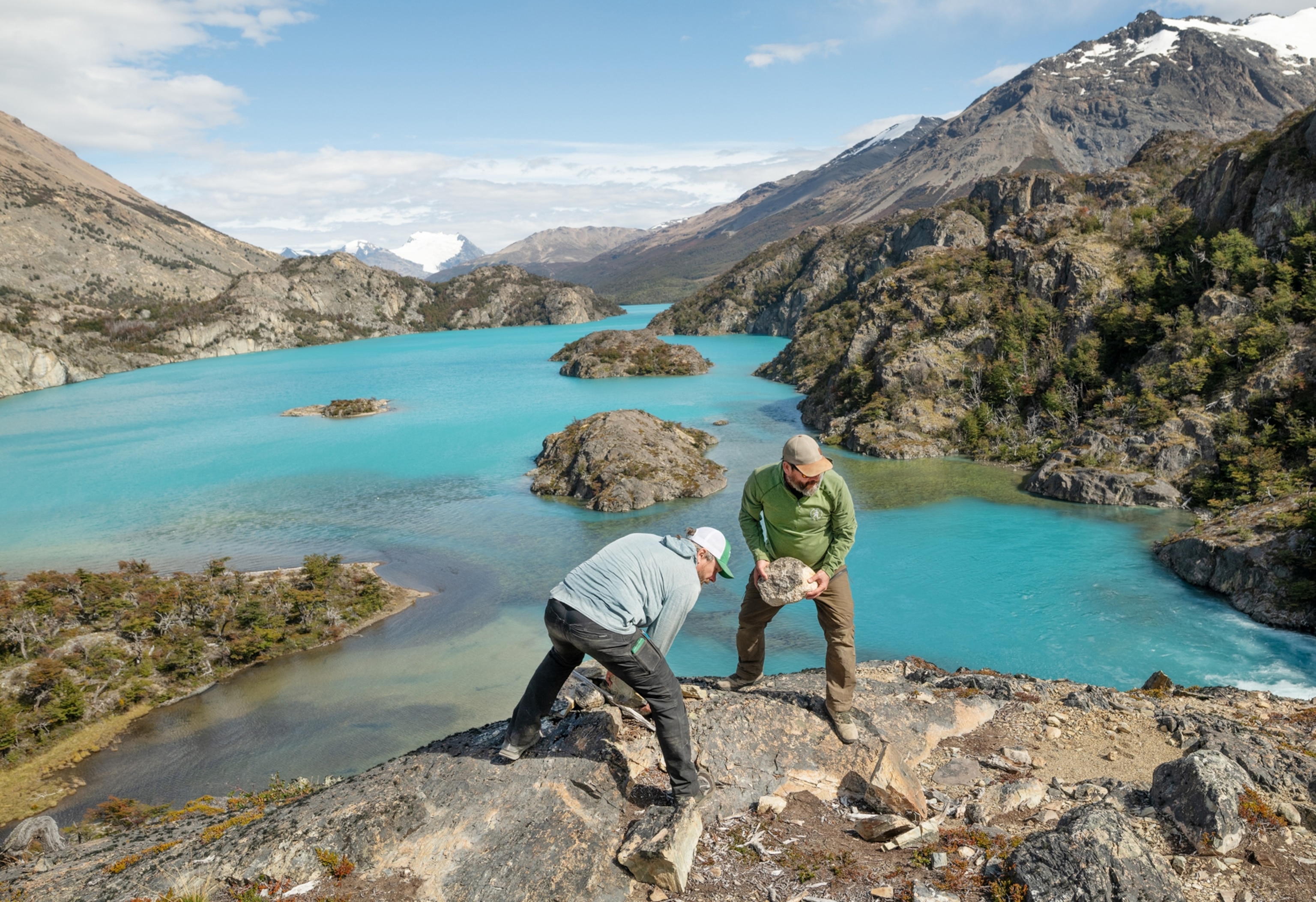 Two men are seen on the edge of a precipice leaning down and picking up rocks.