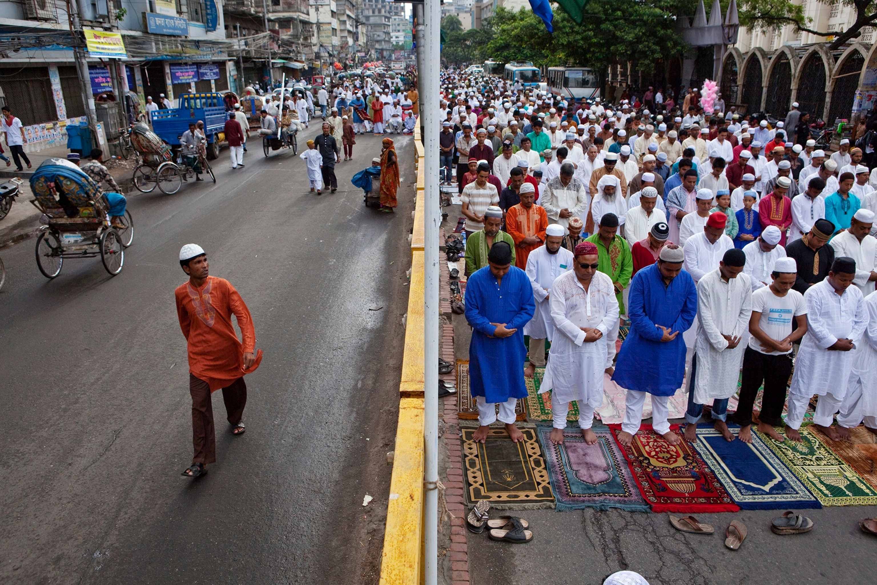 A split street, one side with people walking and the other with a crowd of people standing in front of prayer mats.