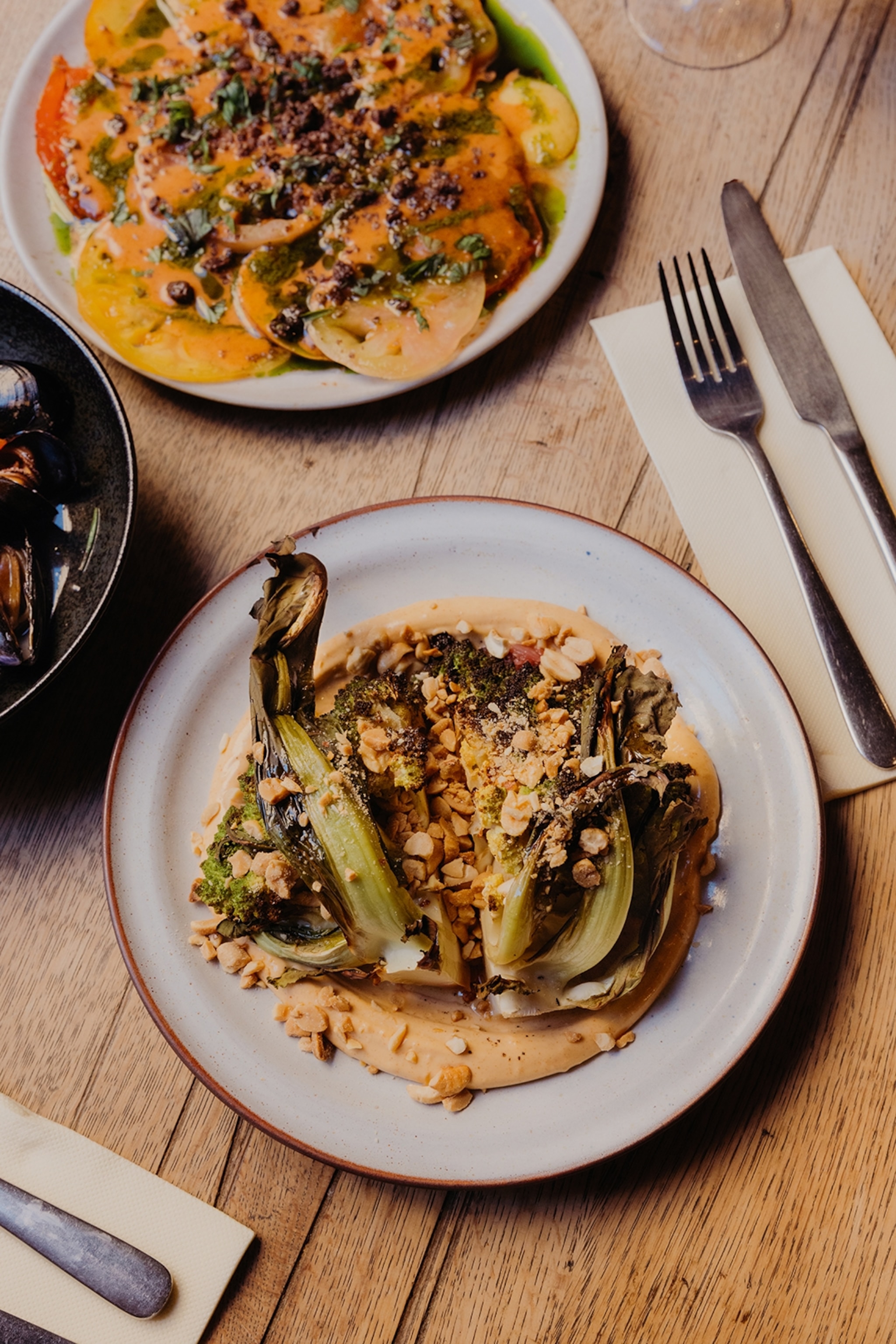 A close-up of a wooden dining table in a restaurant with to plates of plant-based food, including grilled romanesco with cashews and peanuts.
