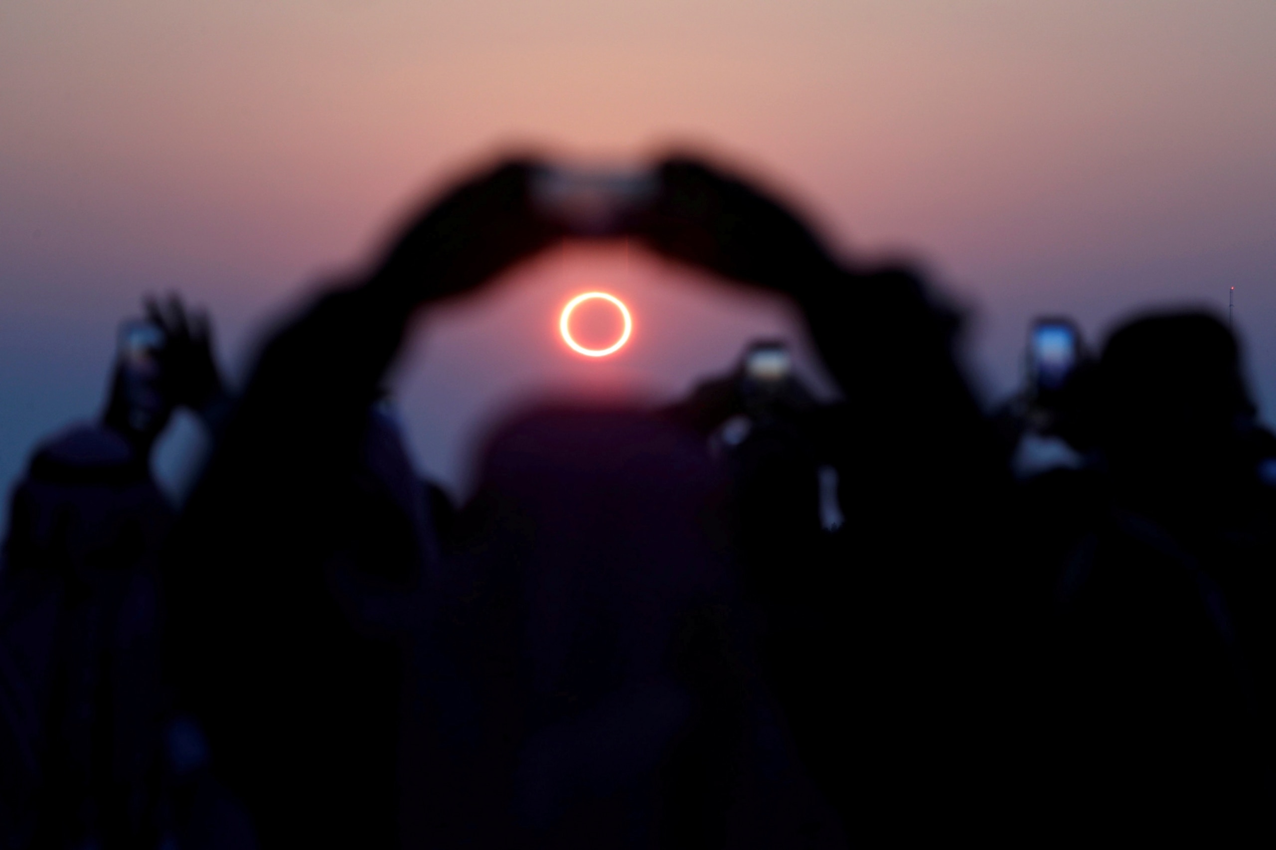 A "ring of fire" eclipse is framed by the arms of a spectator making a photo with their phone.