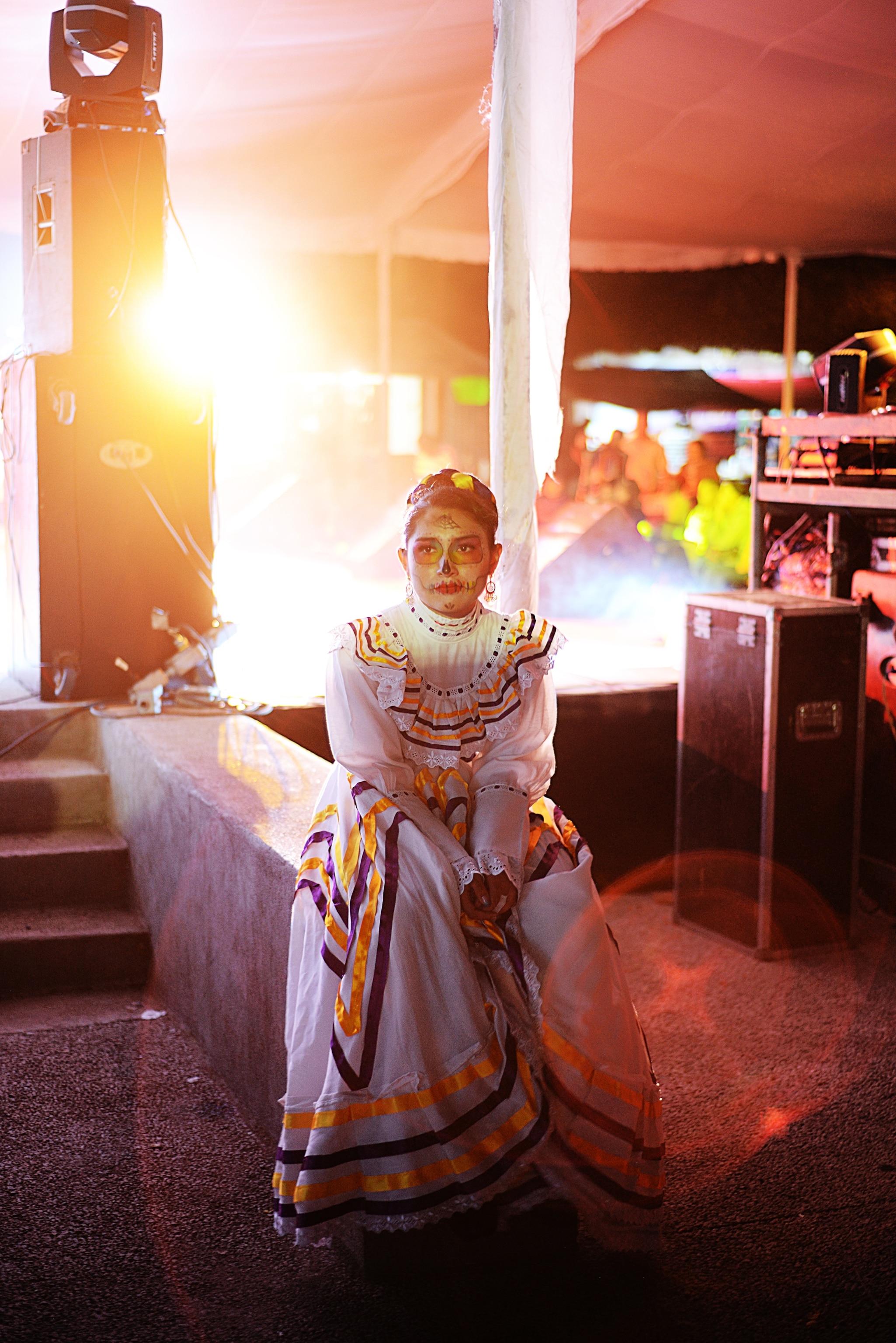 A girl in a white dress with her face painted sits in a room lit by the sun.