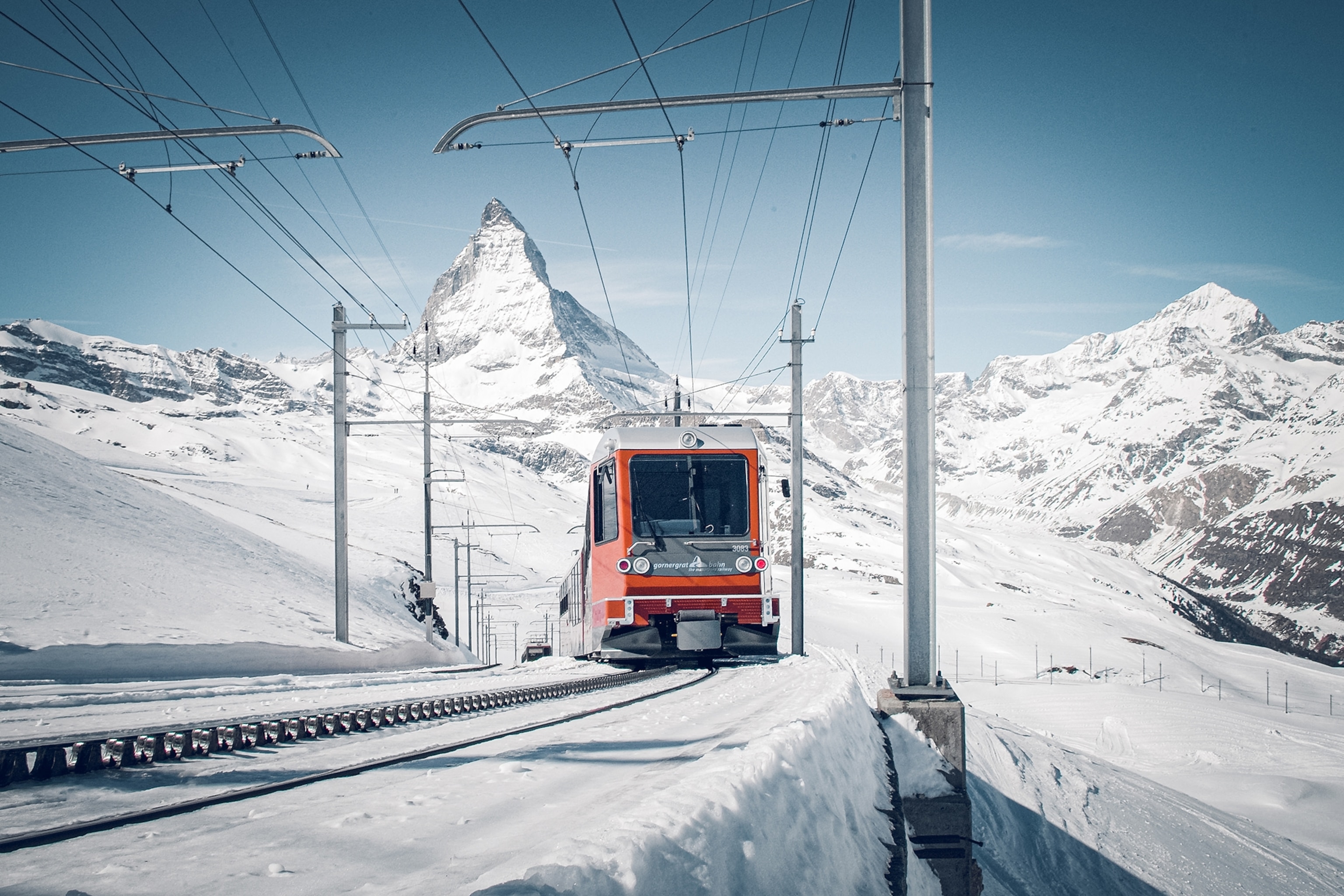 A mountain railway with a train going up a mountain through a snowed in landscape and a mountain peak in the background.