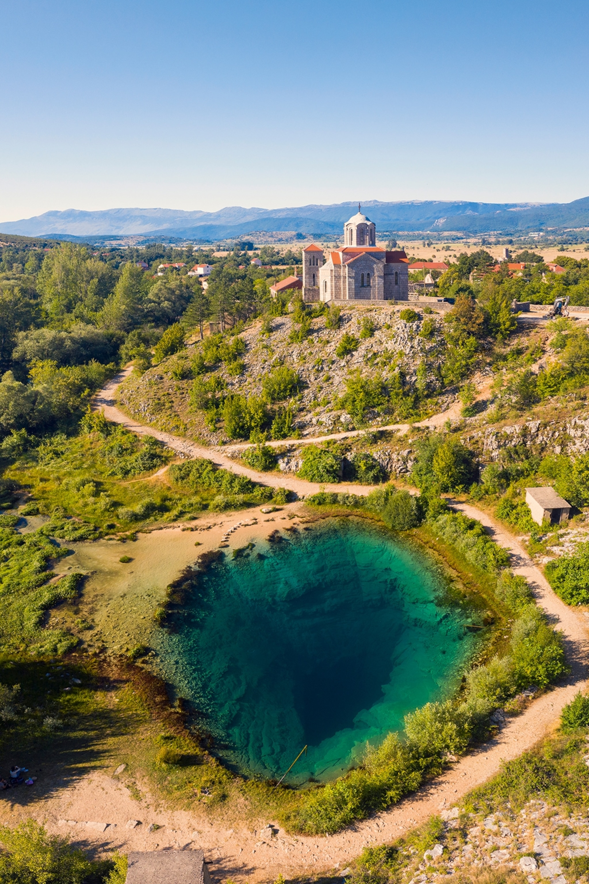 A limekarst landscape with slight hills and falls, a stone church towering over a clear water spring in the shape of a sinkhole.