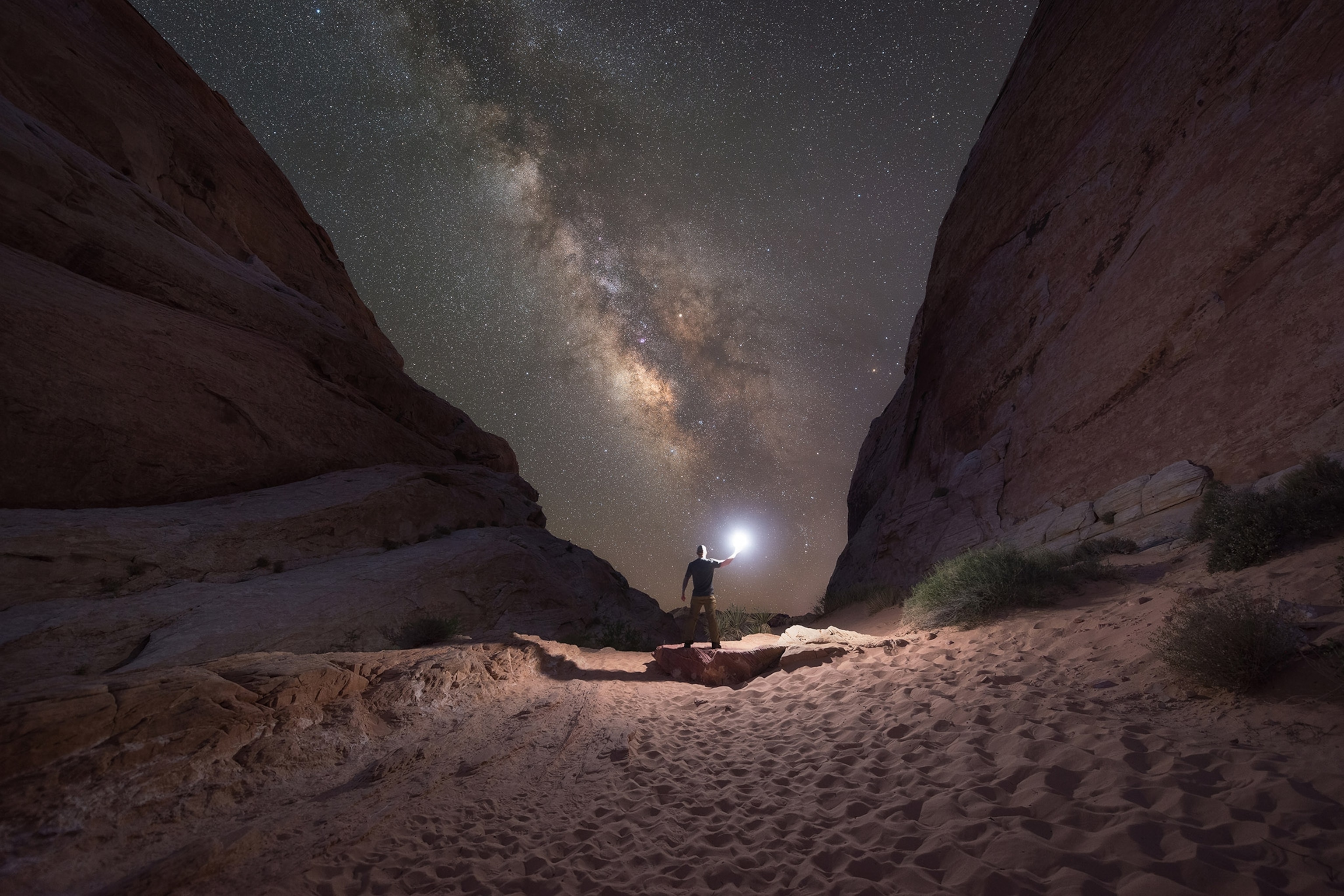 A figure stands at the bottom of a steep gorge, a light in hand illuminating the dark nighttime scene. A bright swath of stars visible in the night sky above.