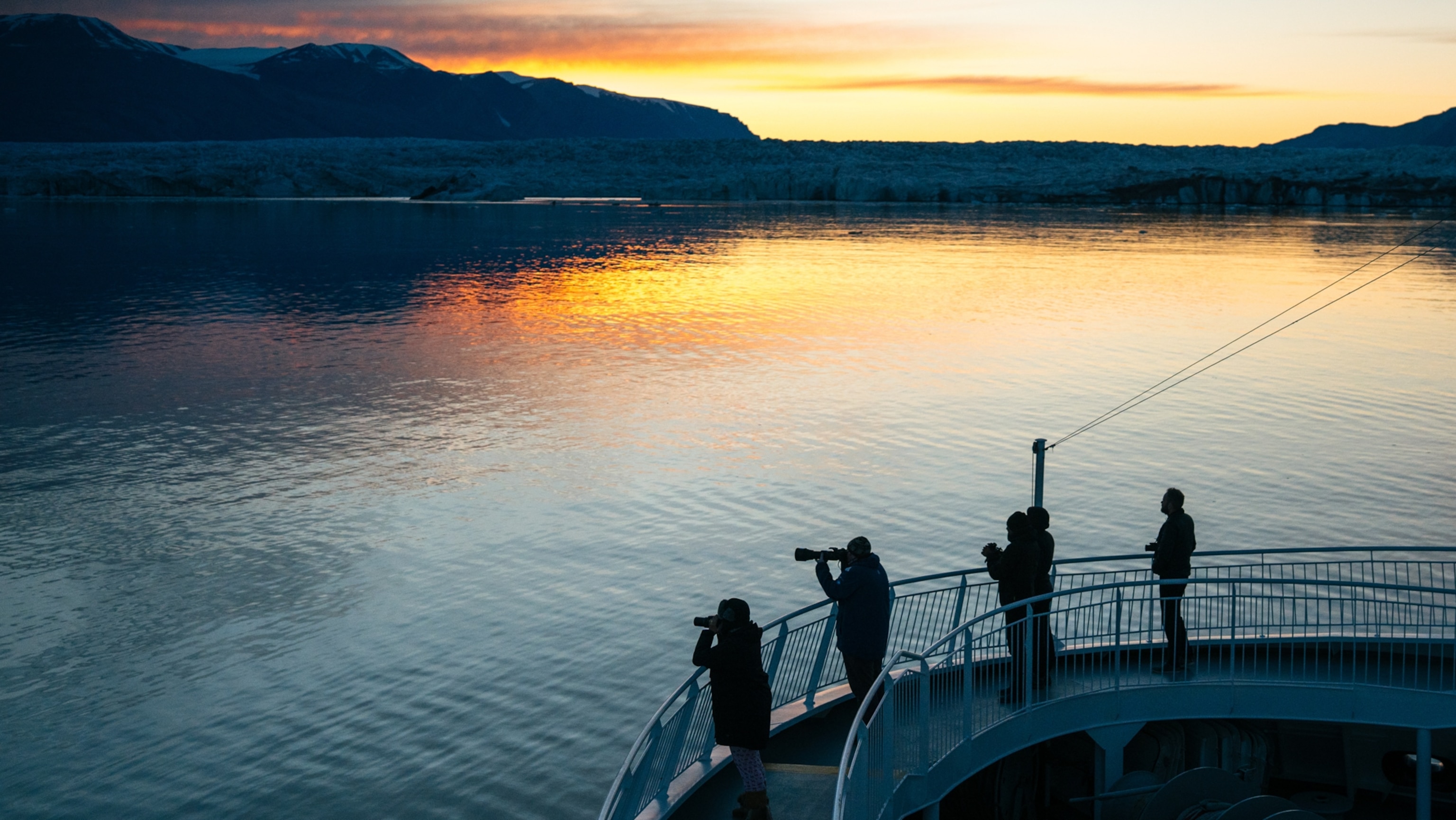 Guests on a cruise ship silhouetted against the icy waters by the edge of a glacier as the sun sets