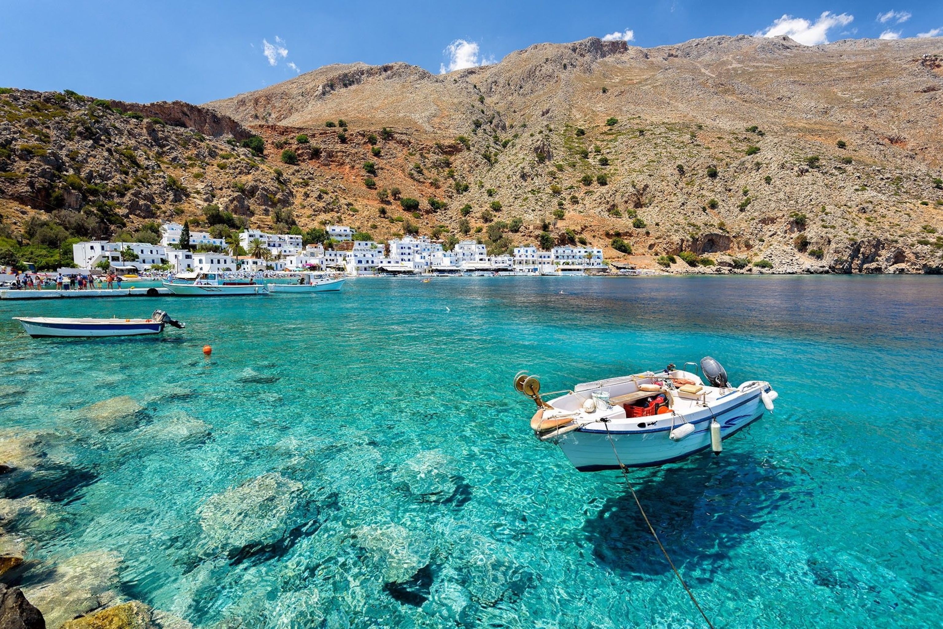A small boat moored in a bay. Loutro is visible in the background. Its buildings are all white.