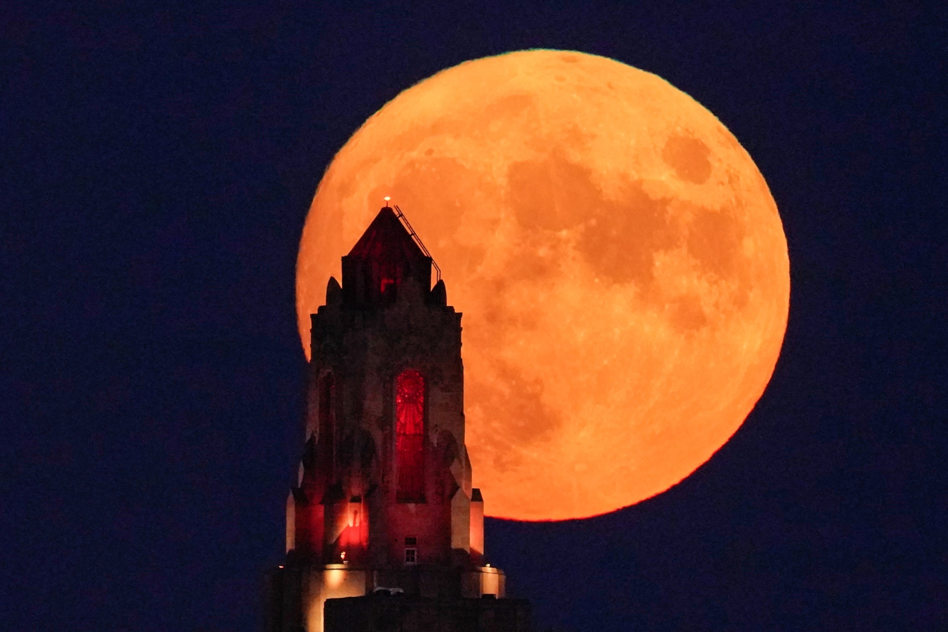 A large, orange full moon rising behind a silhouette of a tower with red-lit windows, set against a deep blue night sky.