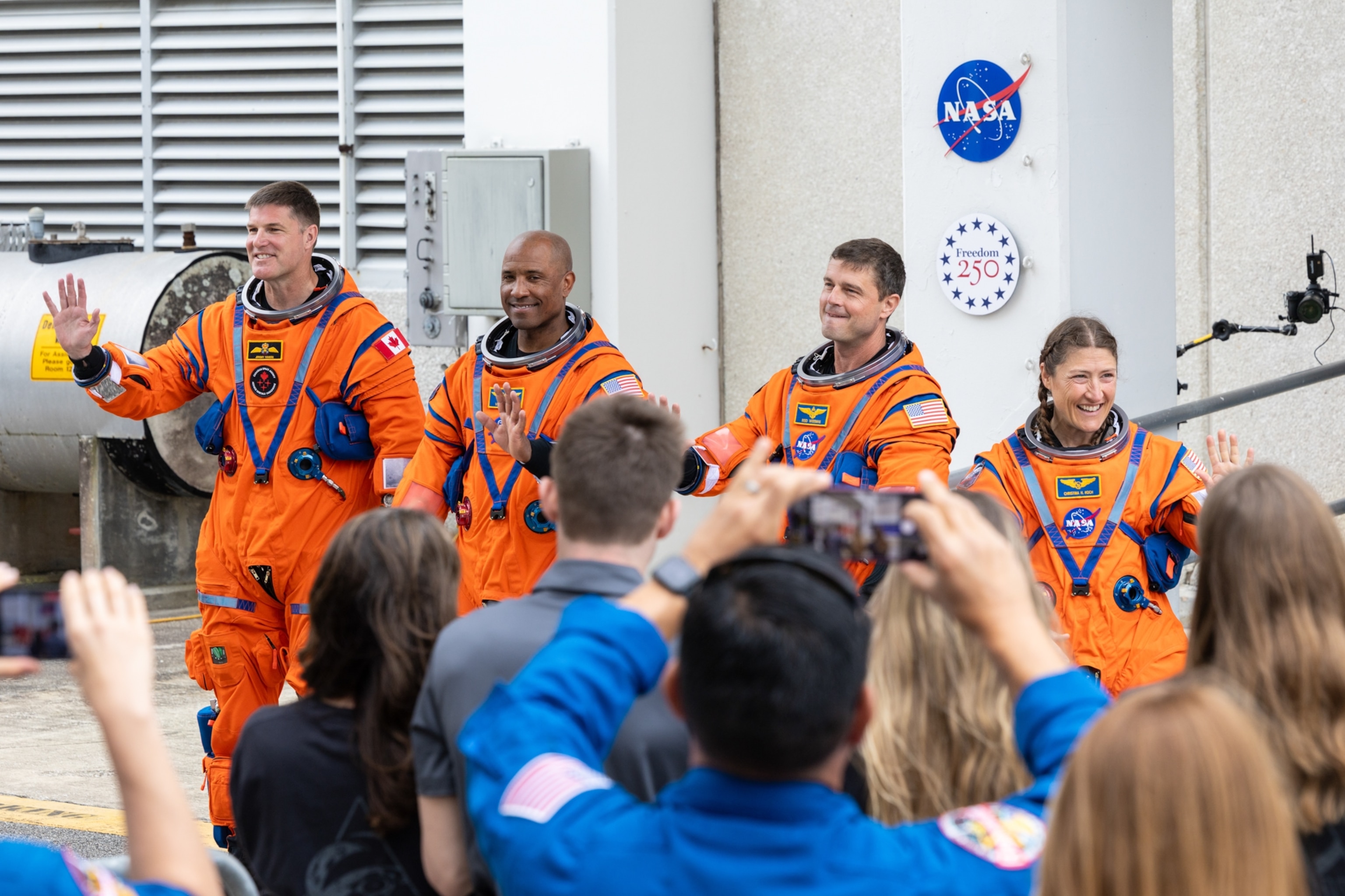 Four astronauts in bright orange space suits wave to an enthusiastic crowd, with NASA signage in the background