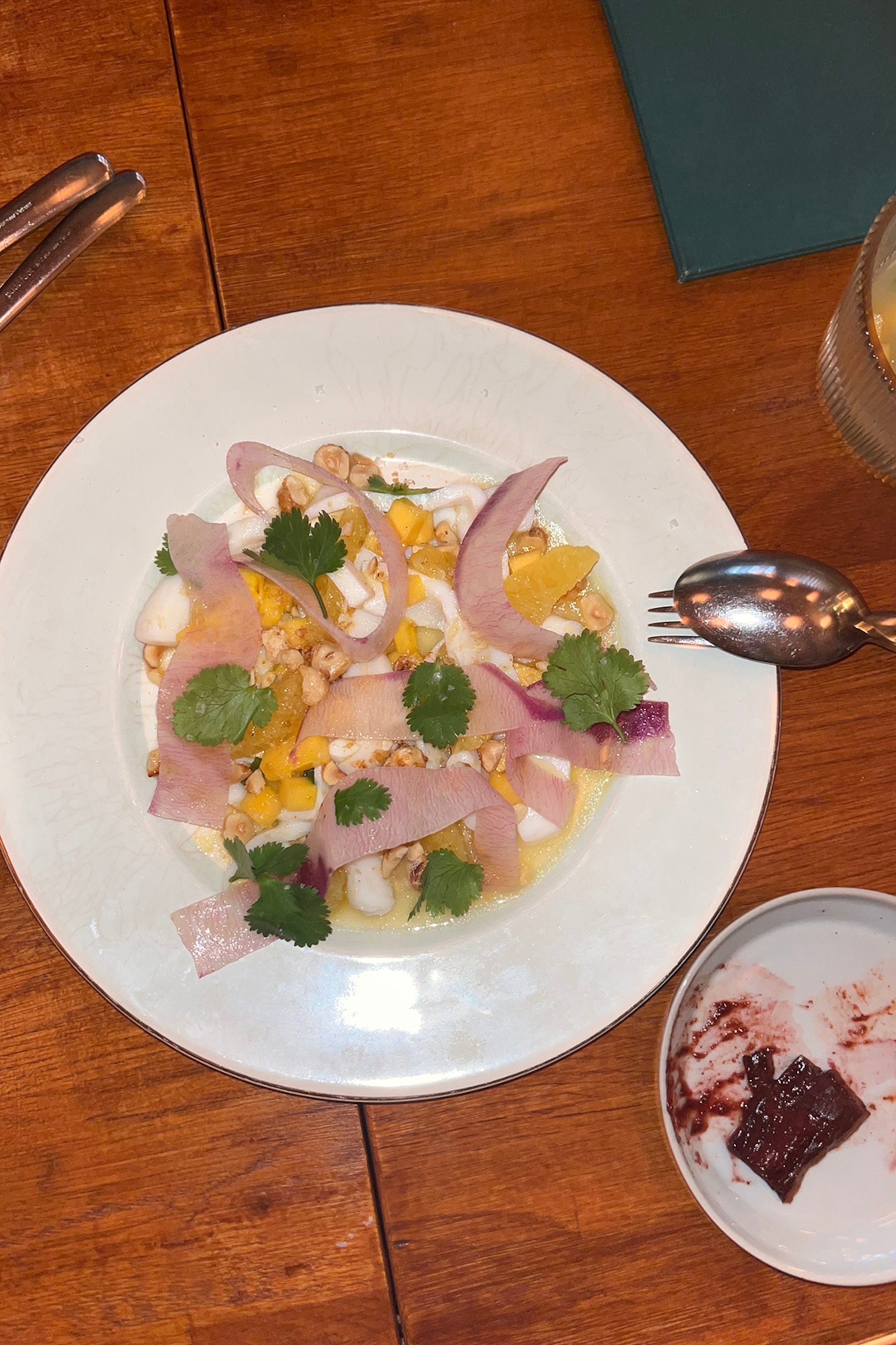 A plate of squid, orange slices, shavings of radish and coriander on a wooden table with cutlery balancing on the rim.