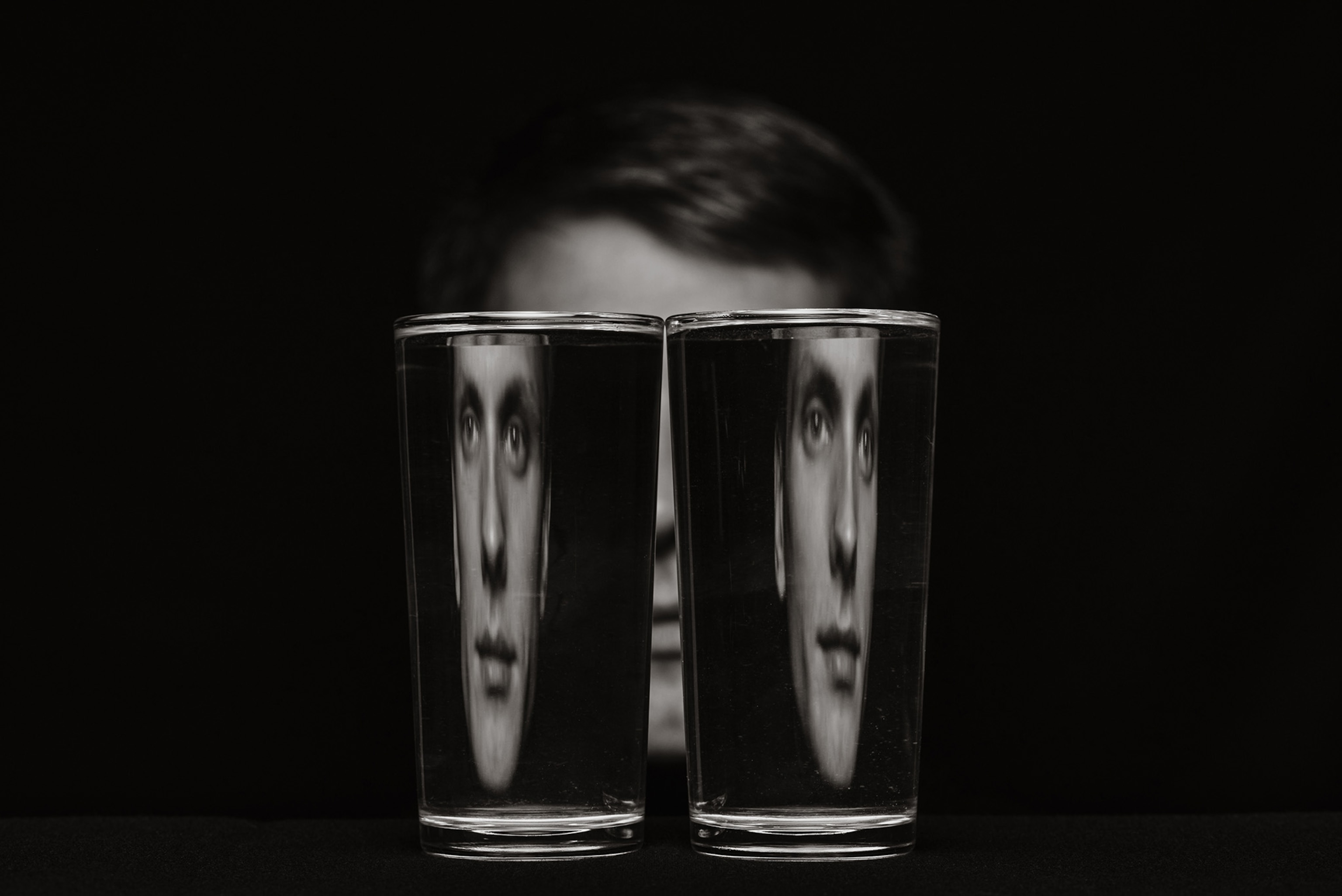 A black and white photo of a mans face refracted through two glasses of water.