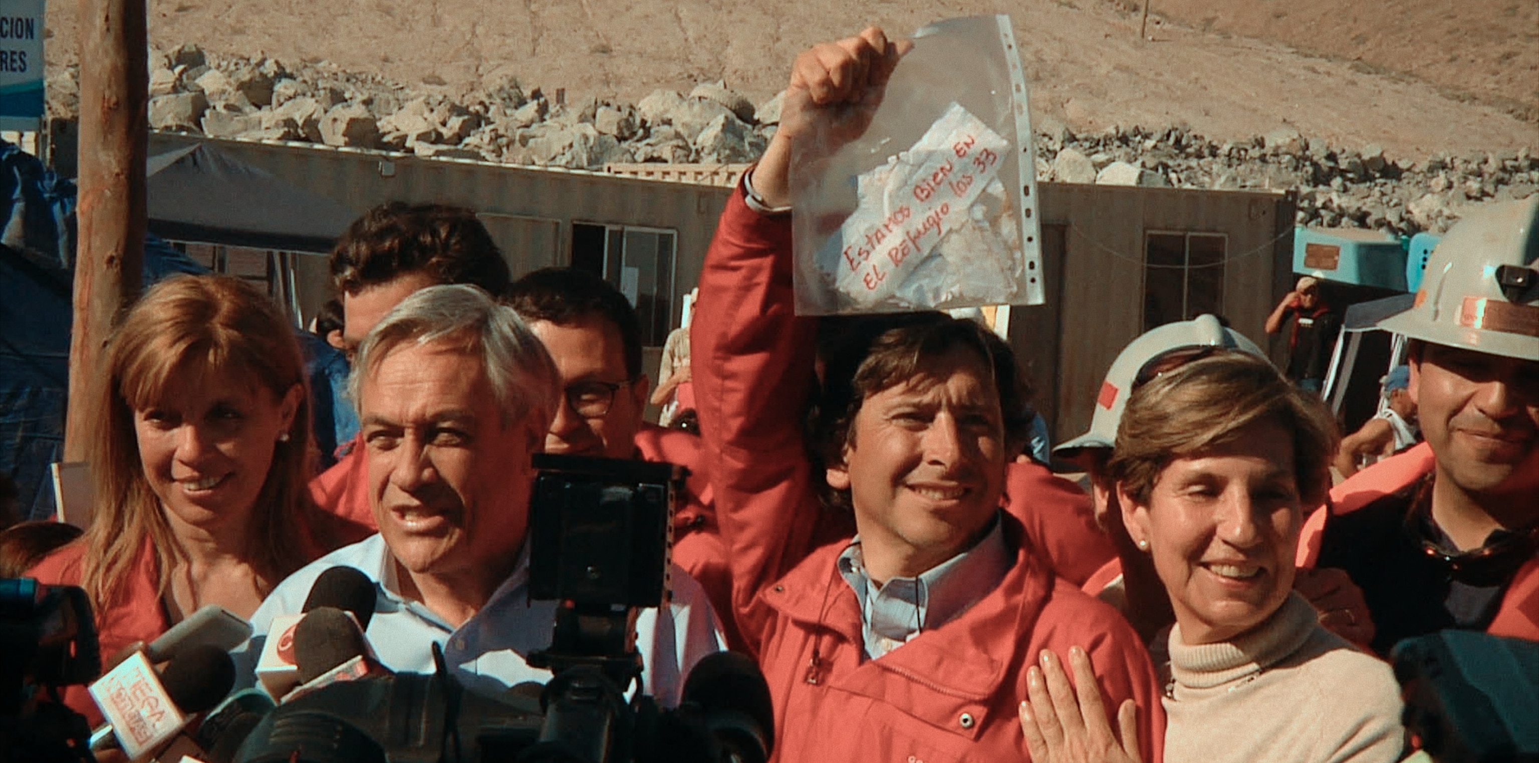 A man speaks into microphones and another man behind him holds up a piece of paper reading "Estamos bien en el Refugio los 33" in a ziplock bag behind the speaker.